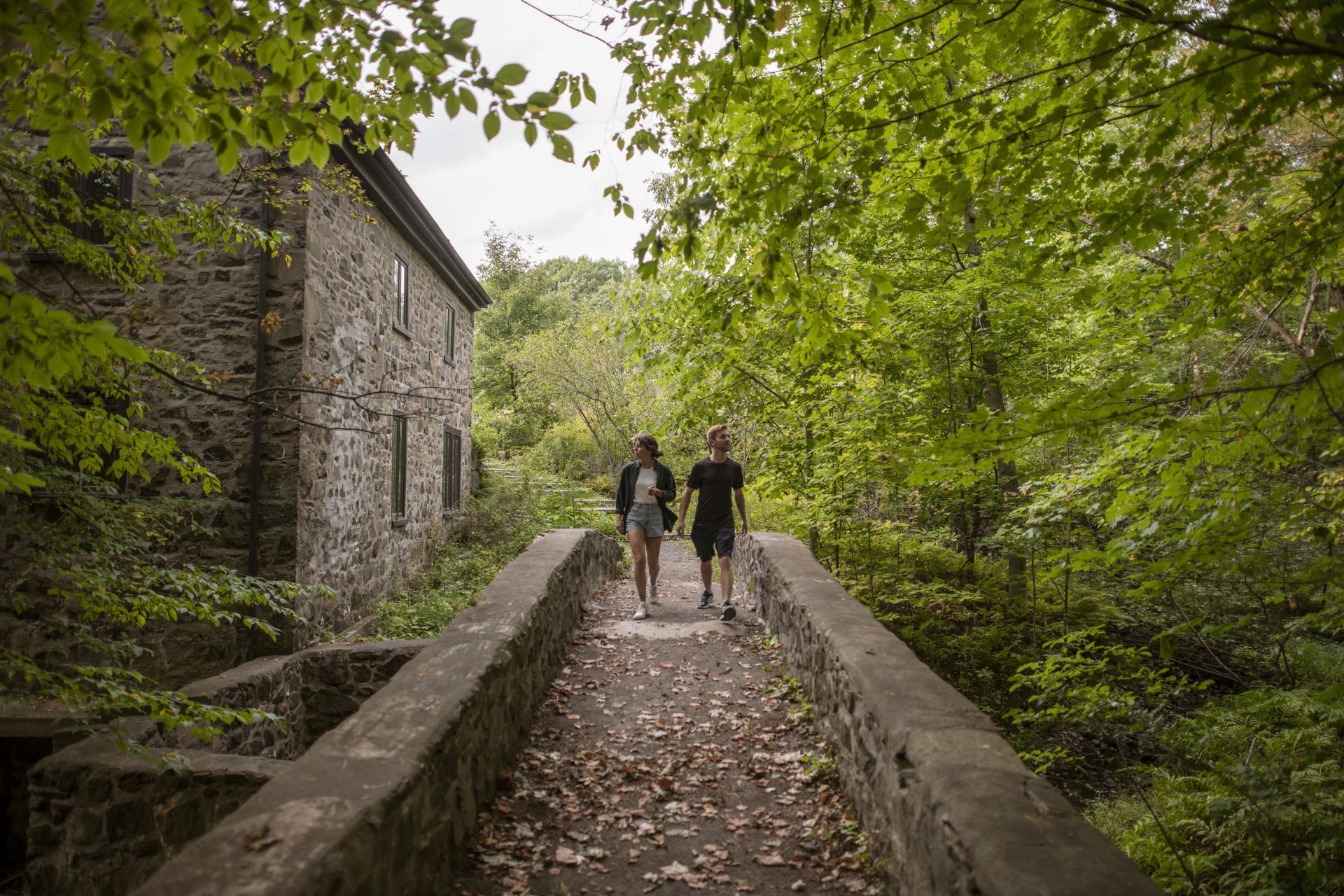 Twee mensen lopen over een stenen brug in Quebec door een lommerrijk bospad langs een oud stenen gebouw.