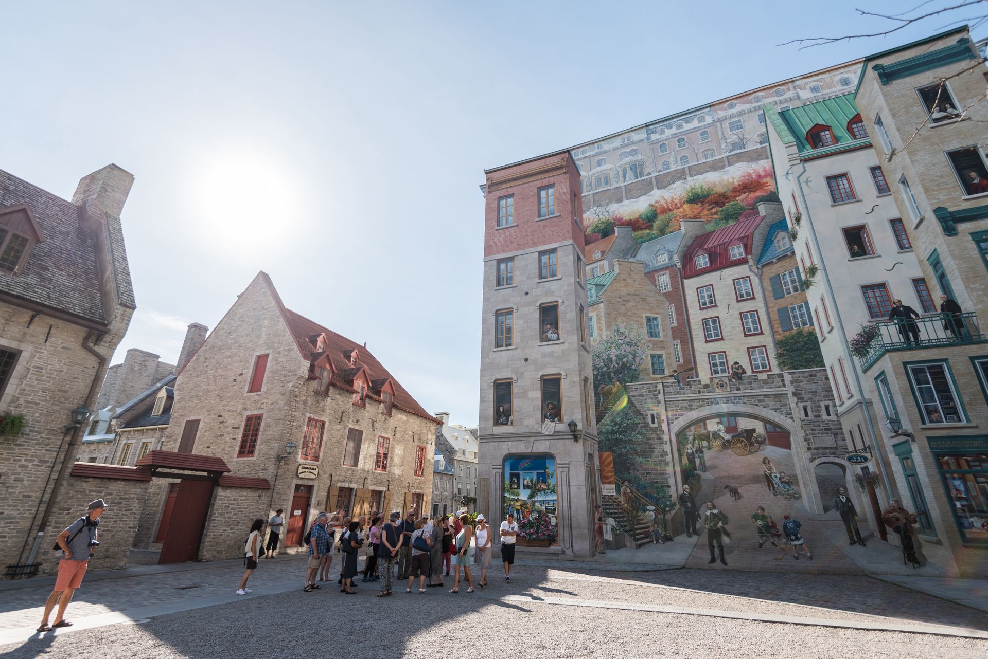 Zonnig geplaveid plein met mensen die zich verzamelen naast kleurrijke historische muurschilderingen op hoge gebouwen van Fresque des Québécois in Quebec.
