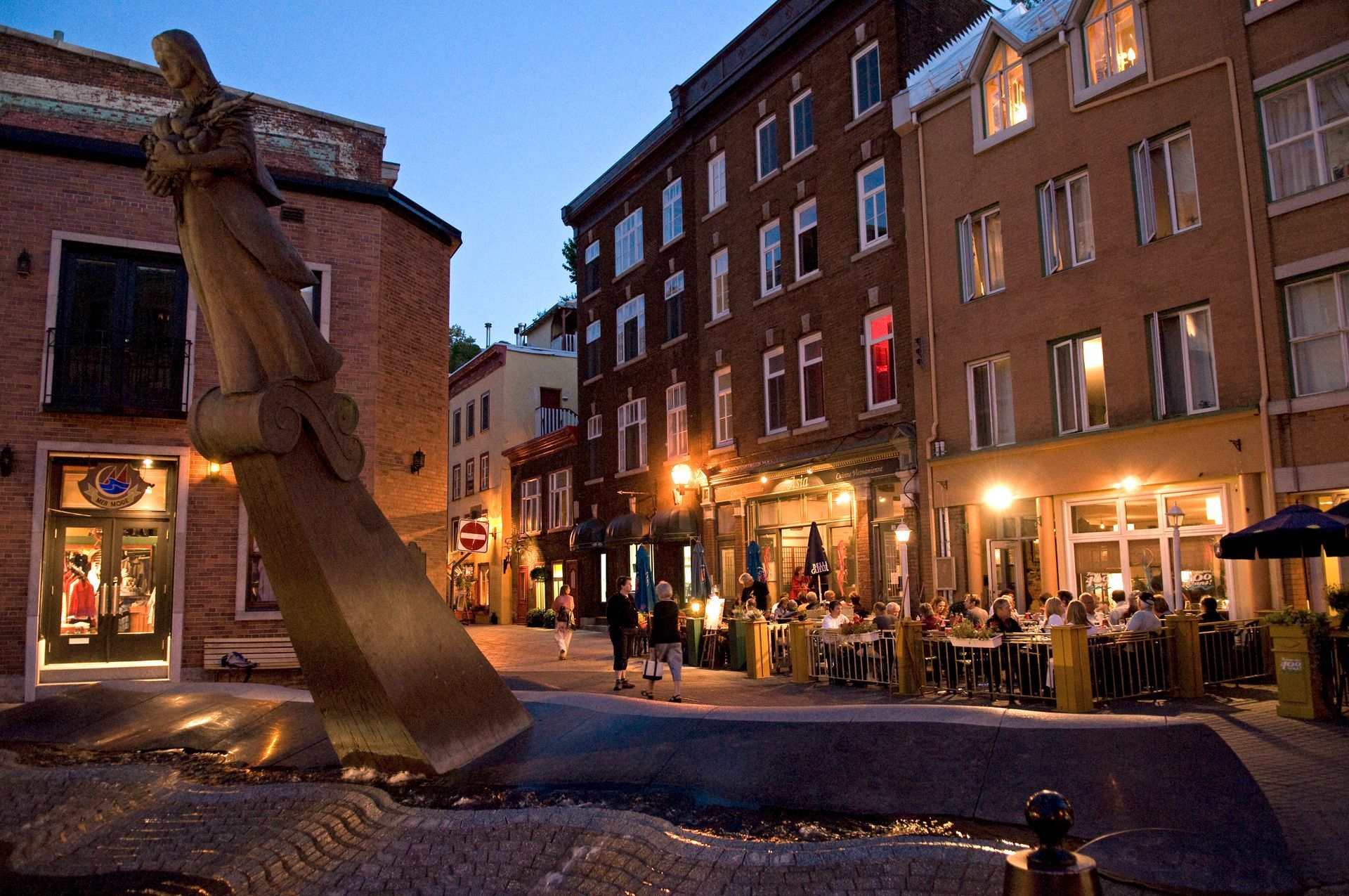 Het stadsplein Place de la FAO in Old Quebec in de avond met een groot abstract beeldhouwwerk, verlichte cafés en bakstenen gebouwen langs de straat.