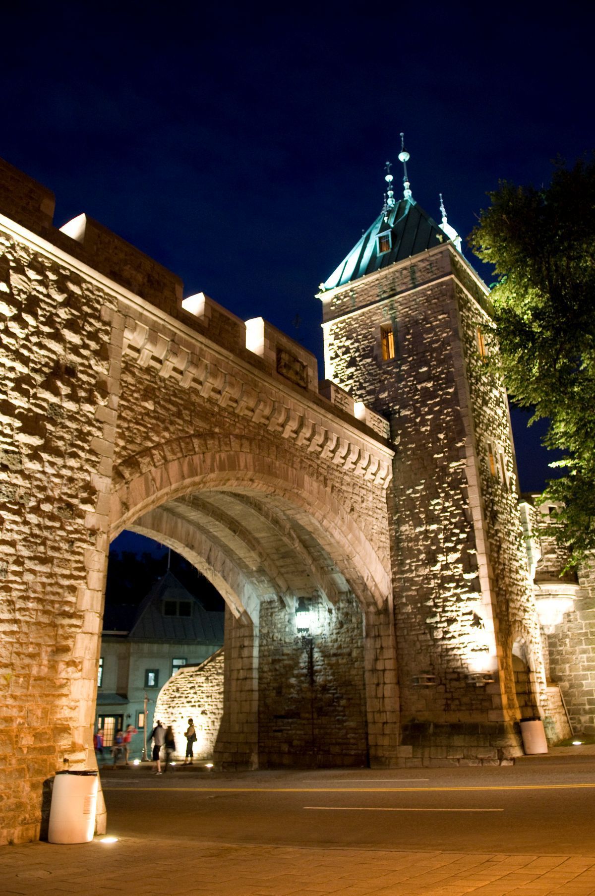De verlichte middeleeuwse stenen poort en toren Porte Saint-Louis in Quebec 's nachts, met een grote boog en een blauw verlichte spits tegen een donkere hemel.