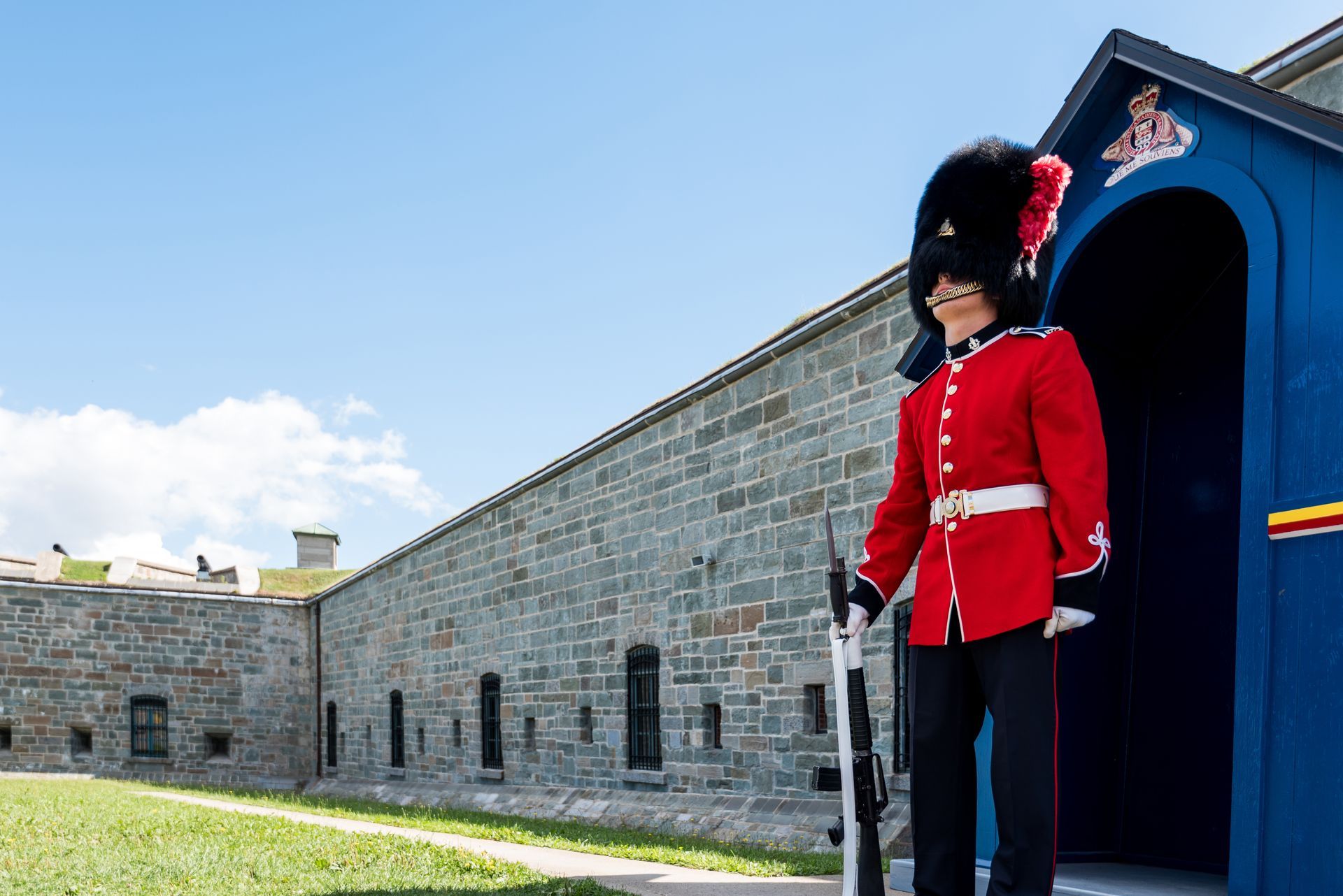 Een bewaker in rood uniform staat naast een blauwe wachttoren bij het stenen fort Citadelle van Quebec onder een heldere hemel.