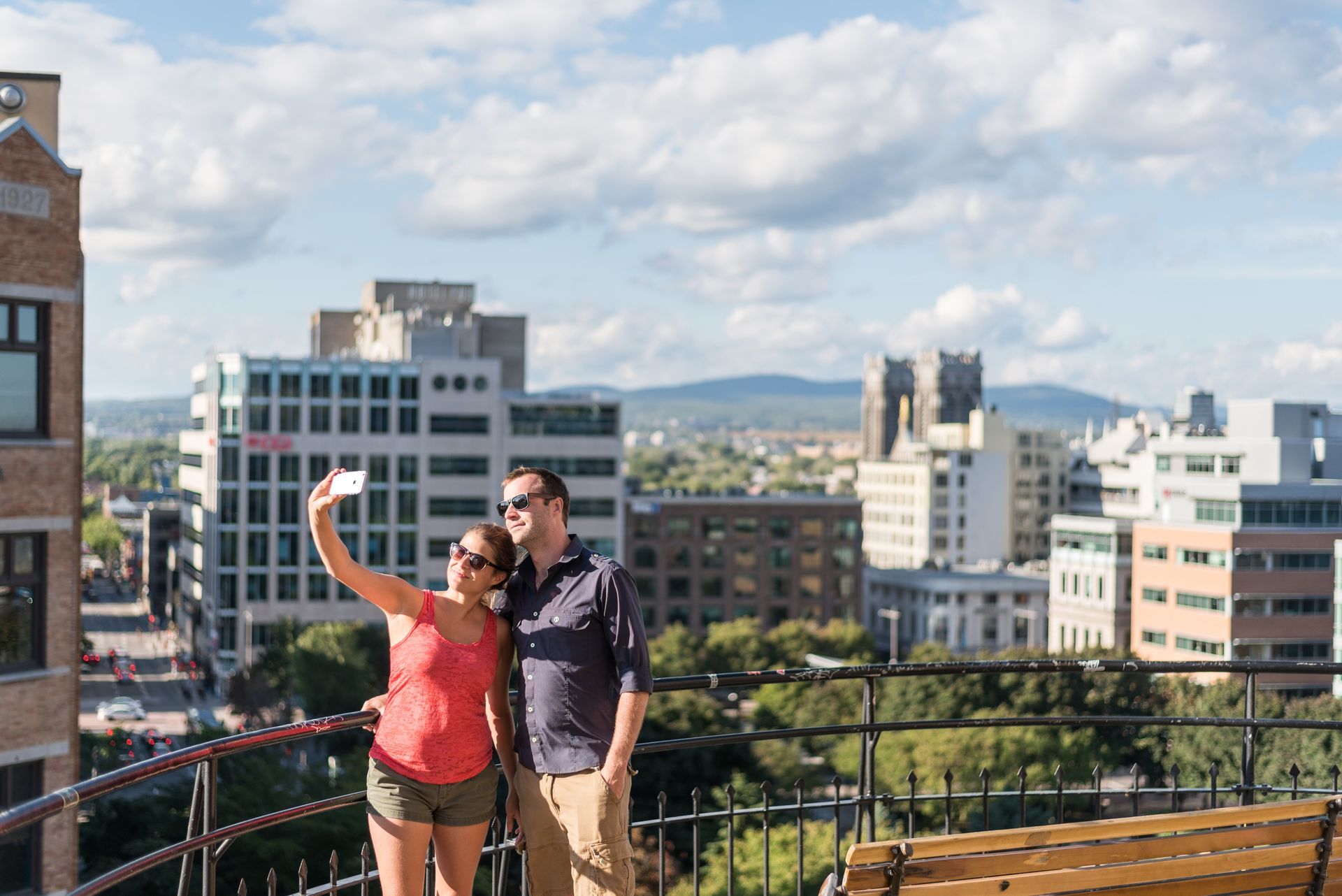 Twee mensen maken een selfie op een dakterras in Quebec met stadsgebouwen en bergen op de achtergrond.