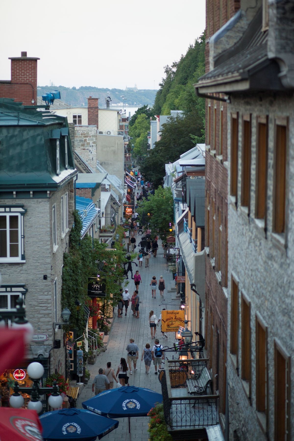 De drukke, smalle straat Quartier Petit-Champlain in Old Quebec tussen stenen gebouwen, van bovenaf gezien, met mensen die lopen en parasols van cafés eronder.