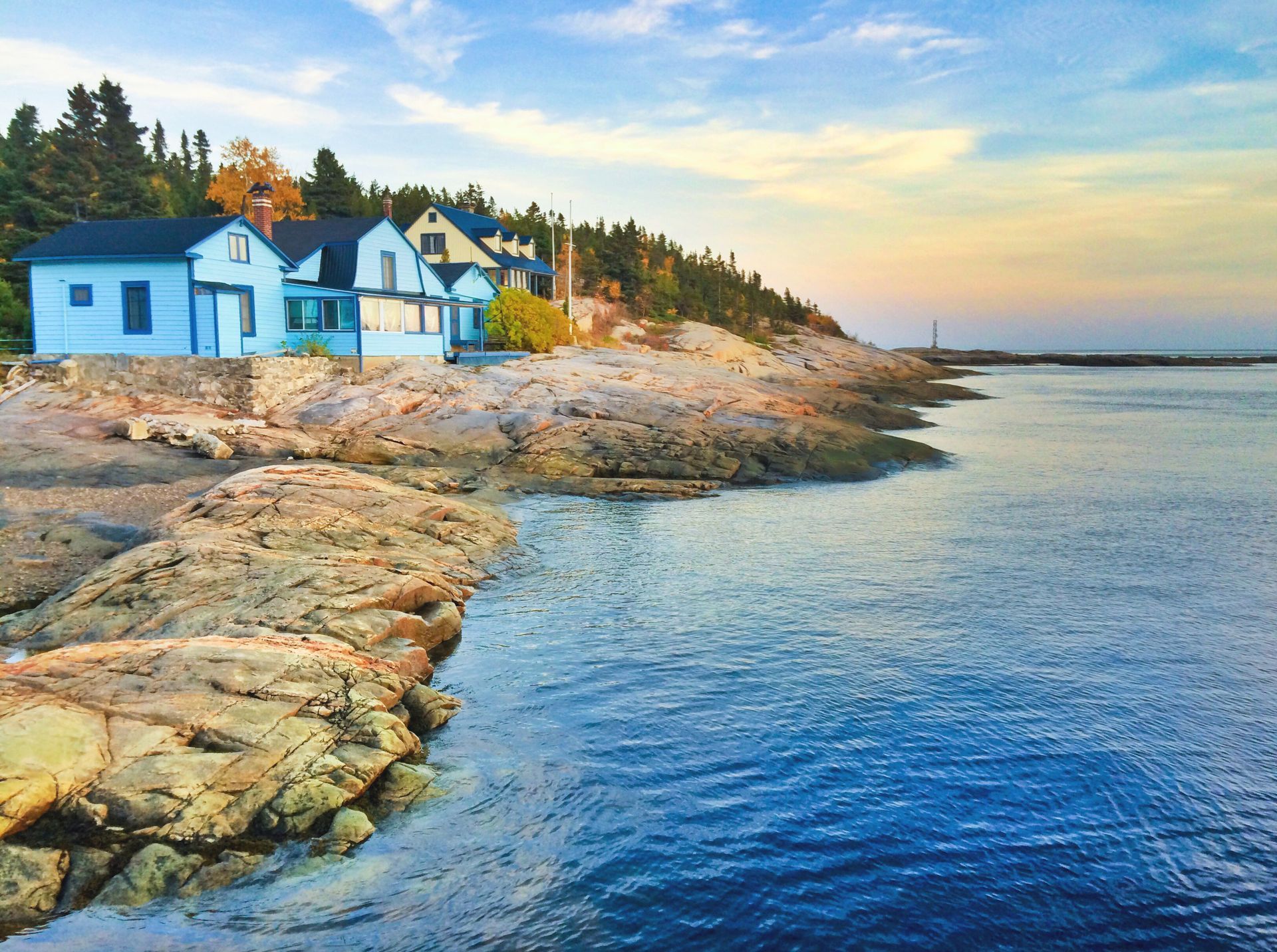 Blauwe huisjes van Tadoussac in Canada aan zee op een rotsachtige kust, aan kalm water bij zonsondergang.