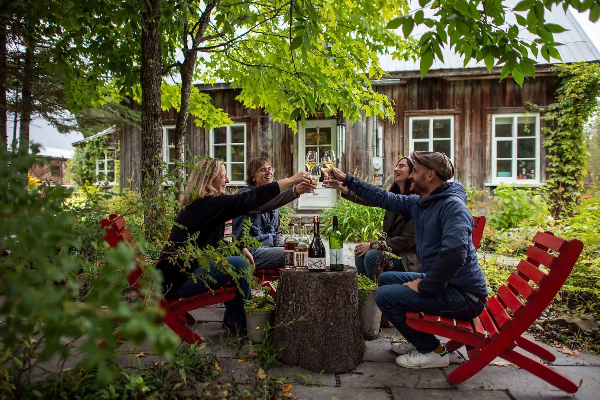 Twee mensen brengen in Quebec, Canada, bij Domaine Vallier Robert een toast uit aan een tuintafel buiten een houten huisje, omgeven door groen.