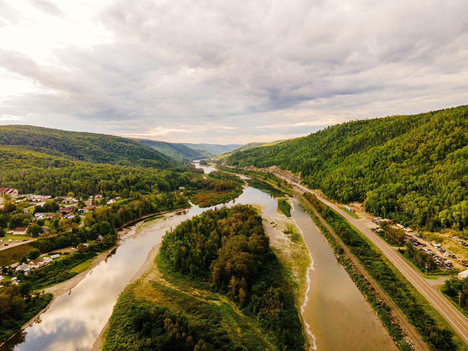 Luchtfoto van de kronkelende rivier Riviere Matapedia in Quabec, Canada, in door groene heuvels onder een bewolkte hemel, met wegen en een klein stadje in de buurt.