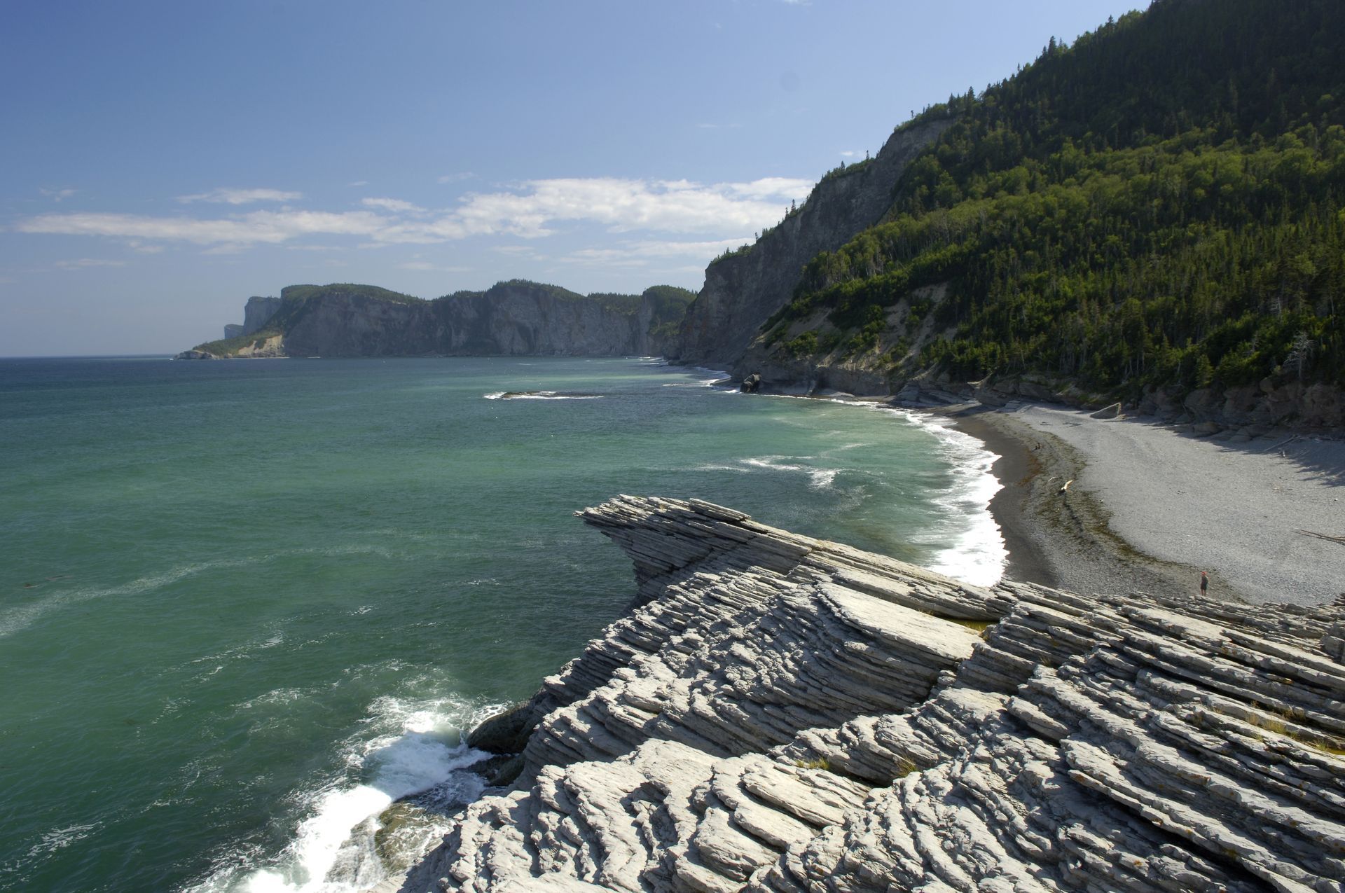 Rotsachtige kustkliffen van het Forillon National Park in Quebec, Canada en een blauwgroene zee met golven langs een gebogen strand onder een heldere hemel.