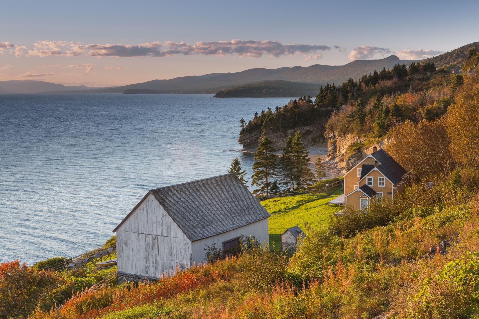 Een heuvel aan de kust in Quebec, Canada, met twee kleine huisjes, herfstbomen en een kalme zee onder een roze hemel.