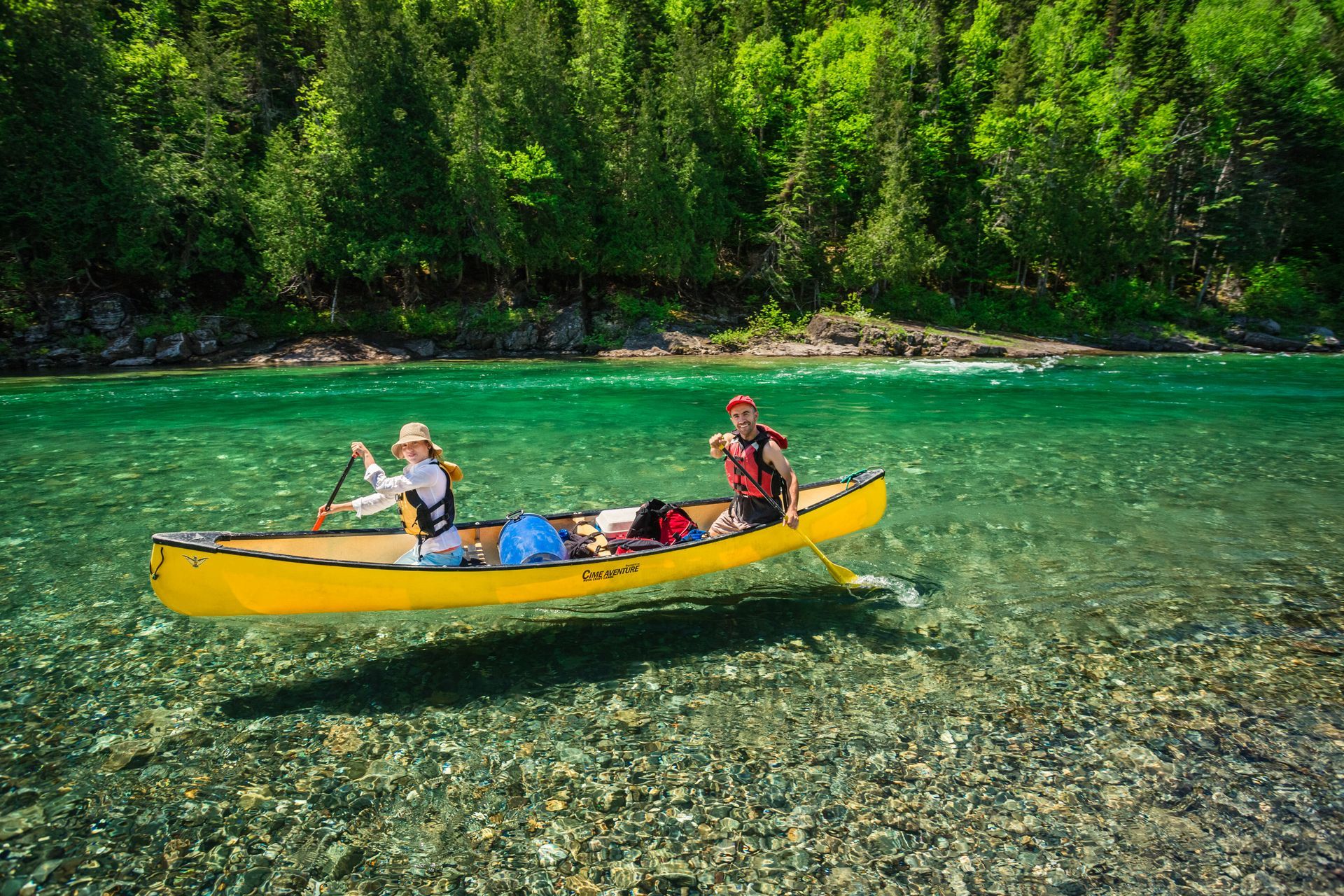 Twee mensen in een gele kano op de heldere rivier Bonaventure in Quebec, Canada,, met een rotsachtige oever en een dicht bos op de achtergrond.