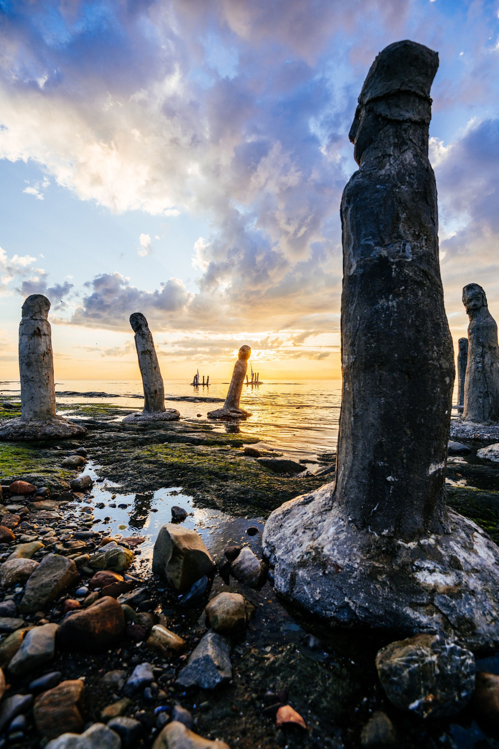 Houten sculpturen n de Saint Lawrence River bij Sainte-Flavie, Quebec Canada, op een rotsachtige kustlijn bij zonsondergang, met golven en een kleurrijke hemel.