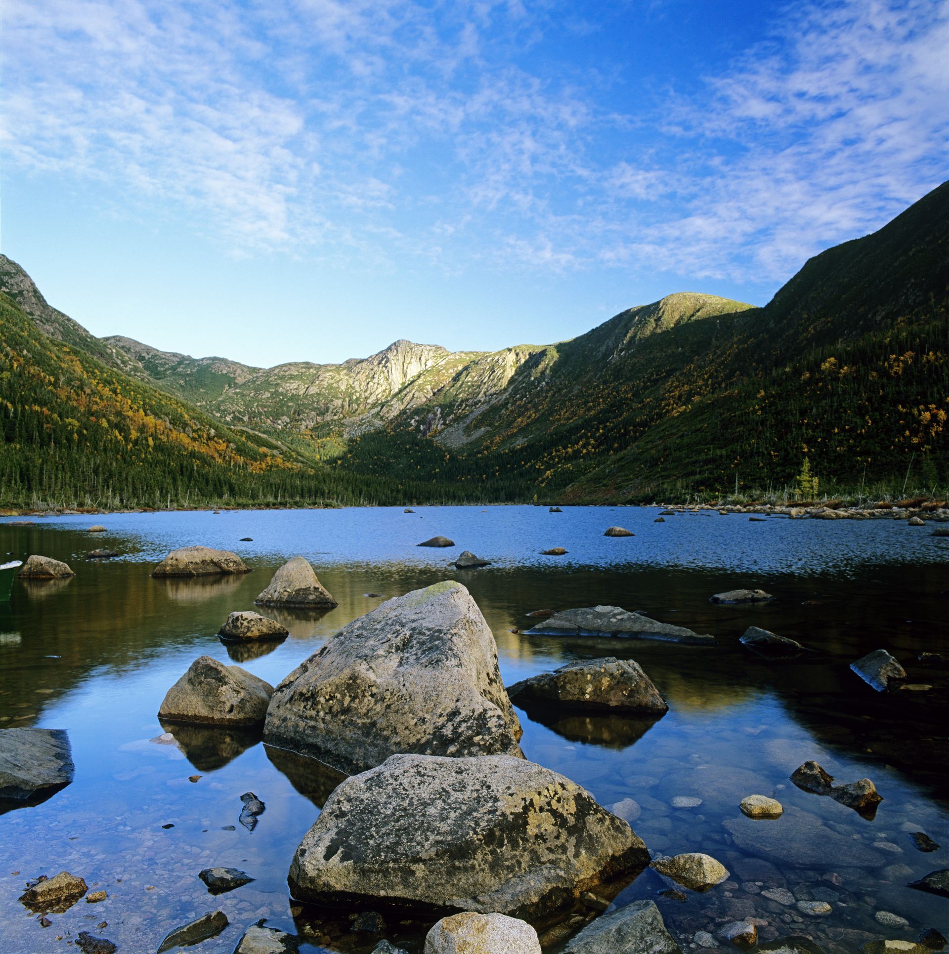 Rotsachtige oever van het meer Lac aux Américains in het Parc national de la Gaspésie met helderblauw water, bergtoppen en een stralend blauwe hemel.