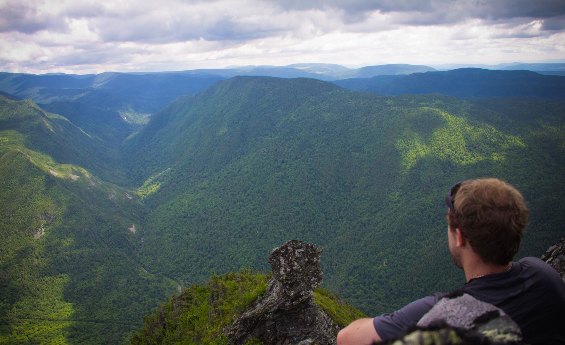 Een persoon vanaf een rotsachtige klif in het Parc national de la Gaspesie, Quebec Canada, en kijkt uit over een groene bergvallei onder een blauwe hemel.