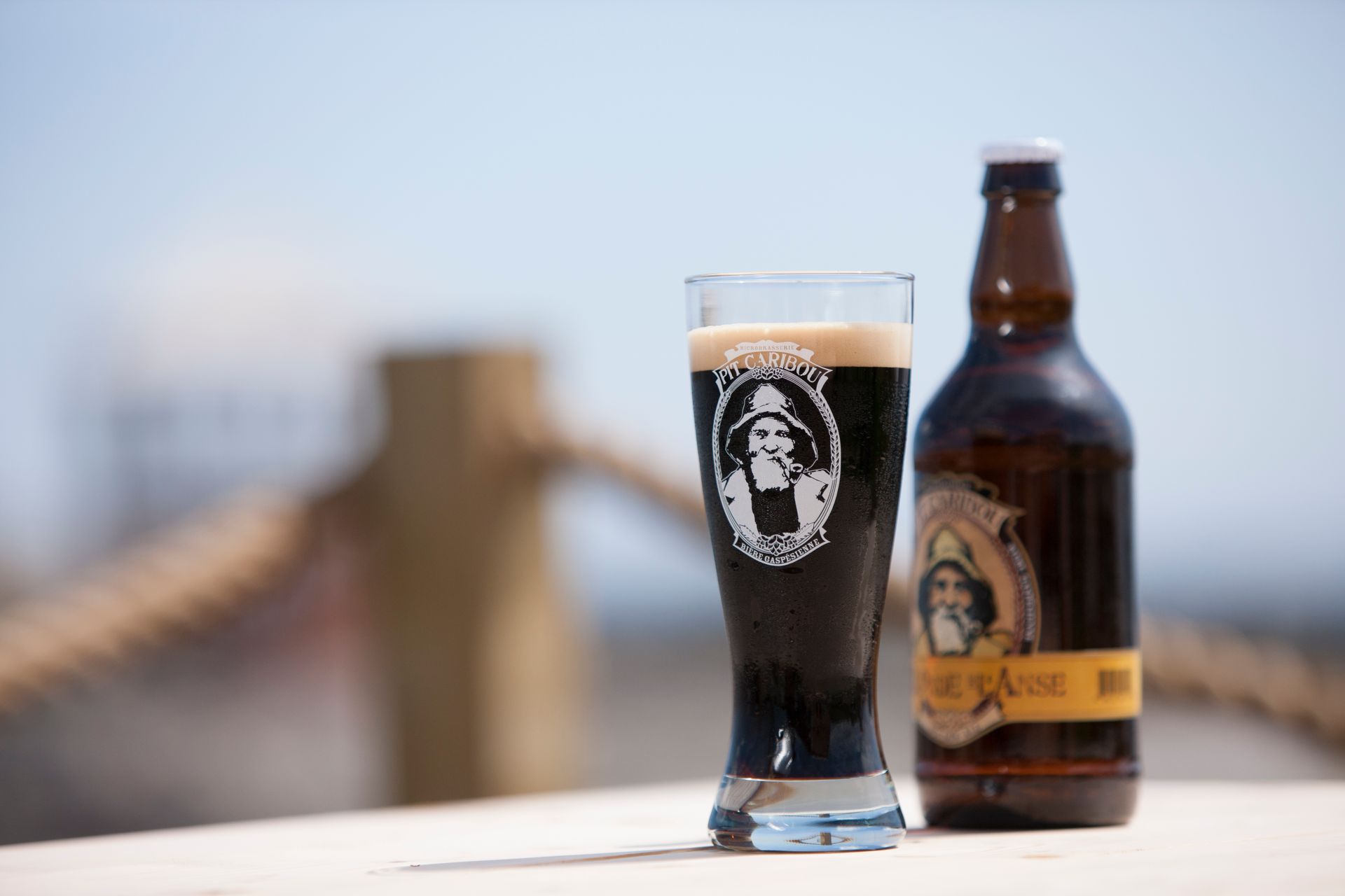 Een bierfles en een gevuld glas op een tafel met een wazige brug op de achtergrond.
