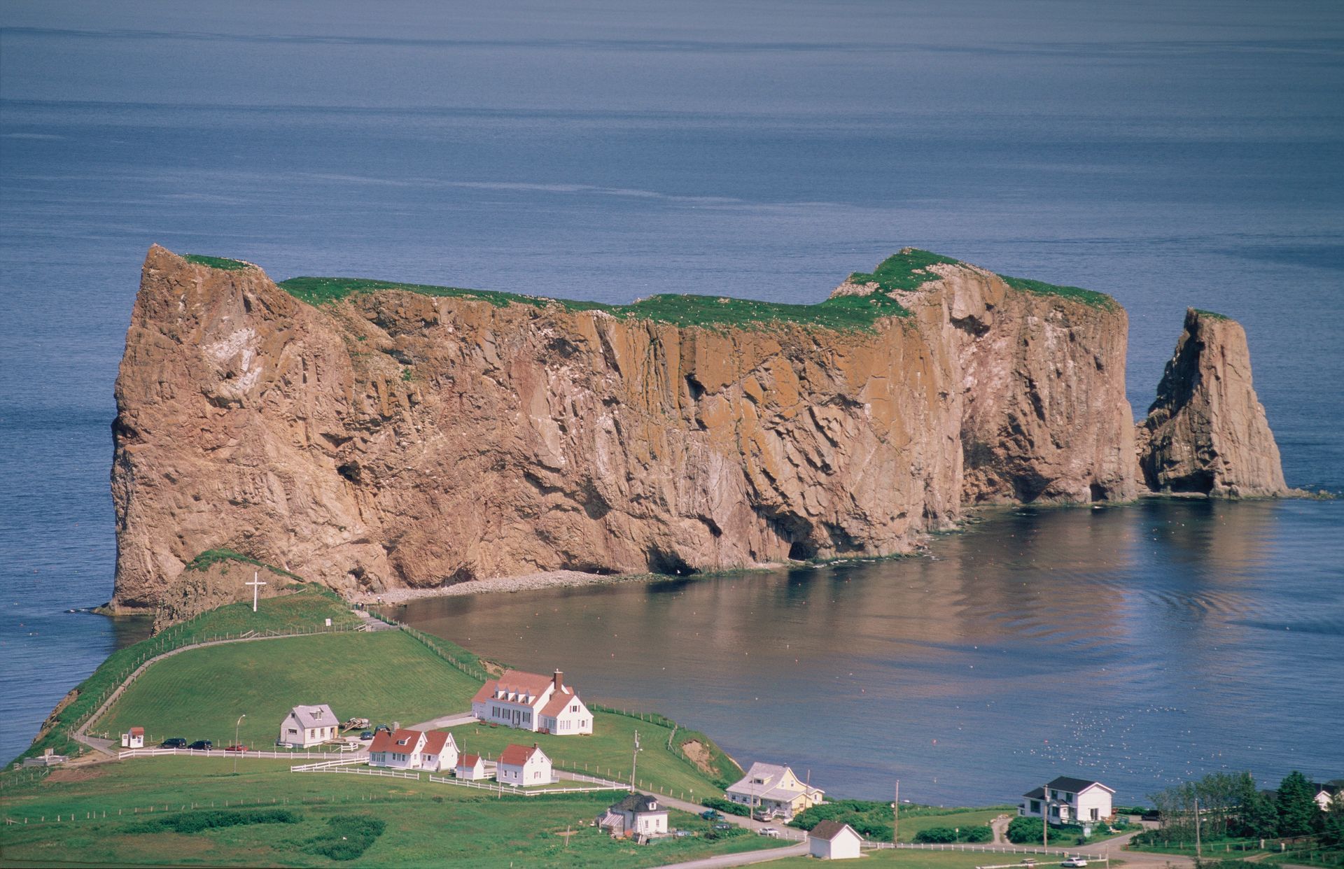 Perce Rock in Canada aan de kust dat oprijst uit de blauwe zee, met groene velden en kleine huisjes op de voorgrond.