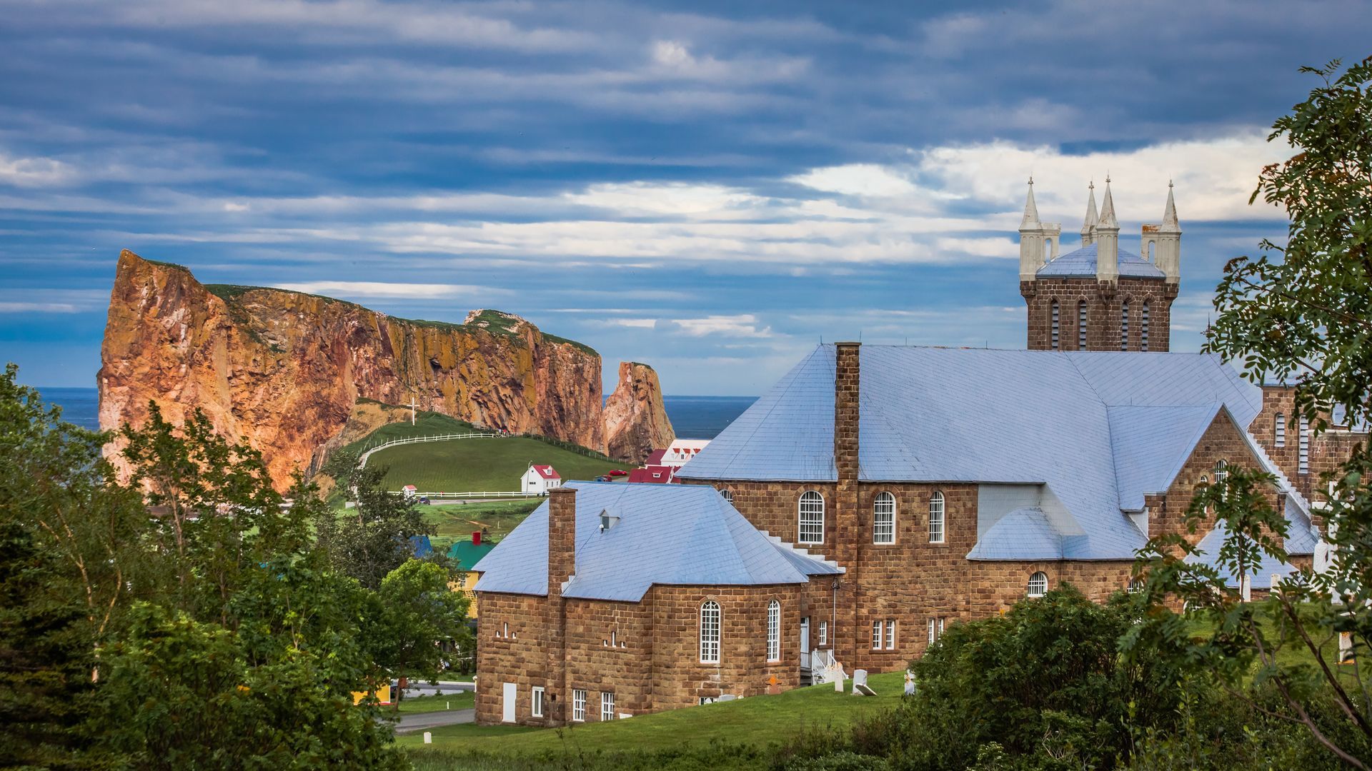 Stenen kerk met blauwe daken voor Perce Rock in Canada onder een bewolkte hemel.