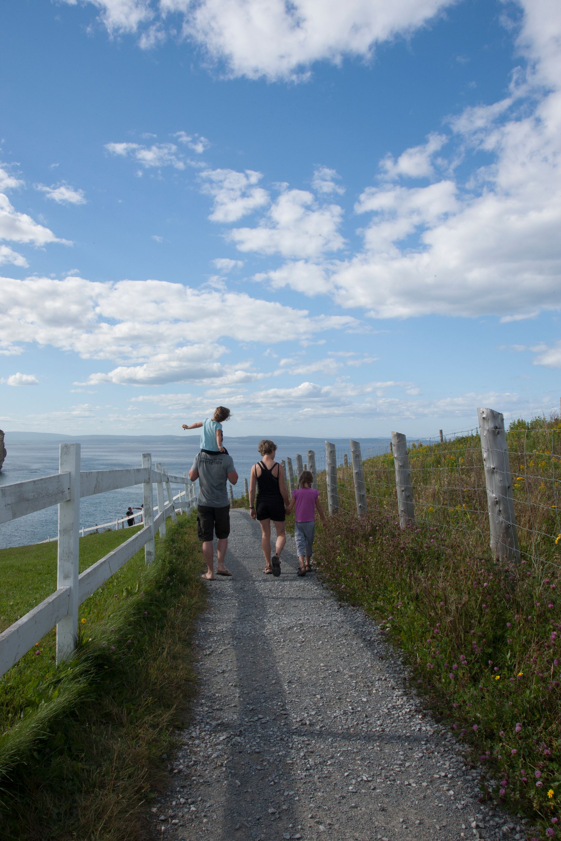 Twee mensen lopen over een grindpad in Perce, Canada, langs een wit hek onder een blauwe hemel met wolken.
