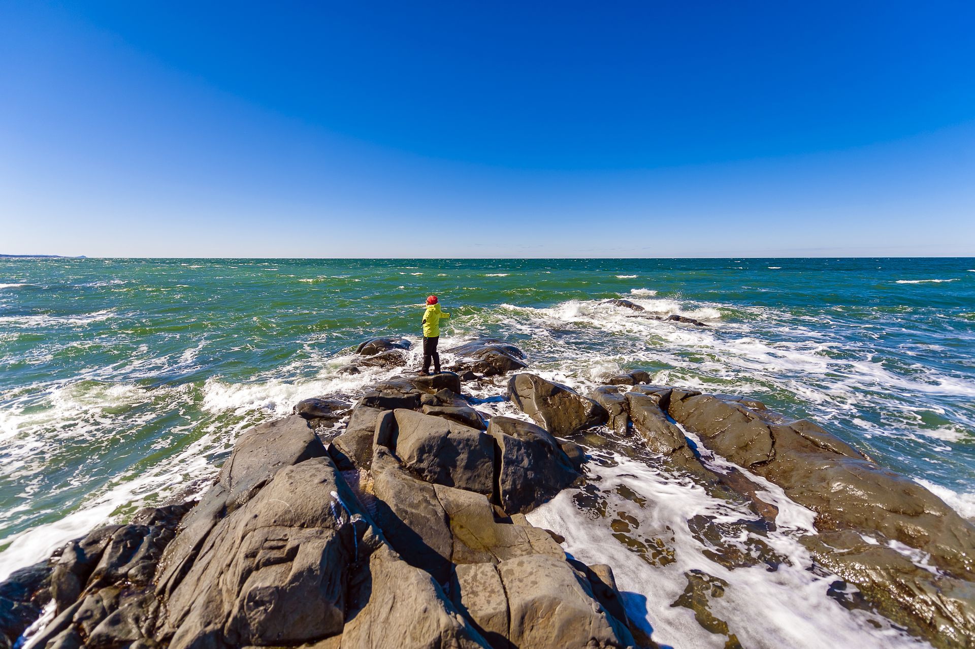 Een persoon staat op een rotsachtige kust in Quebec, Canada, naast een ruige oceaan onder een helderblauwe hemel.