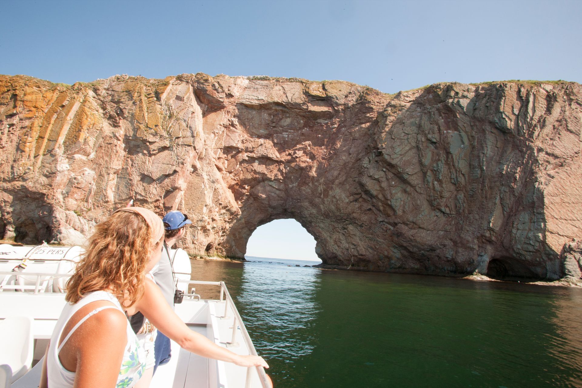 Mensen in een boot die onder de natuurlijke boog van Percé Rock doorvaren, boven kalm groen water.