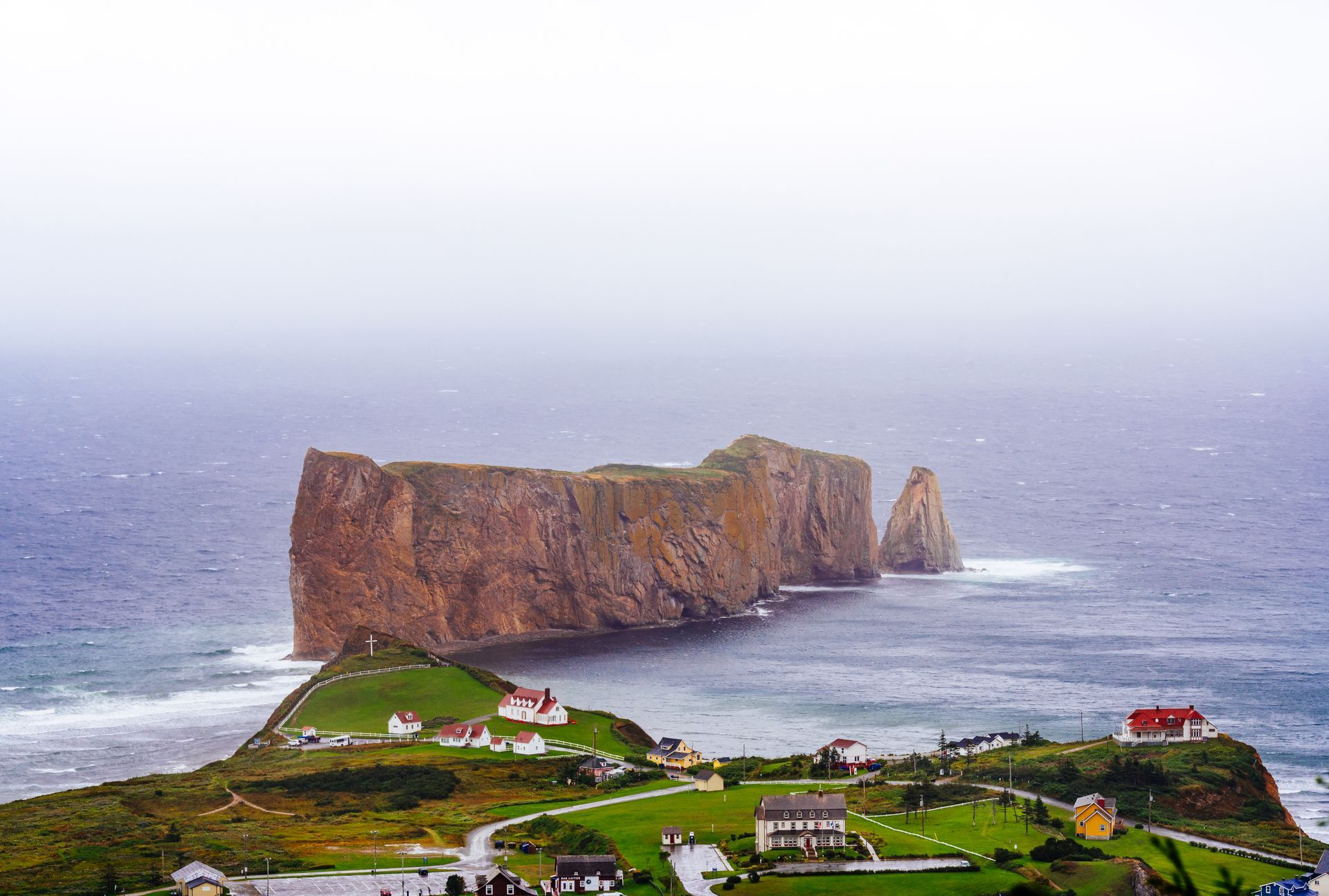 Percé Rock in Quebec, Canada, met huizen op de voorgrond, onder een mistige hemel aan de oceaan.