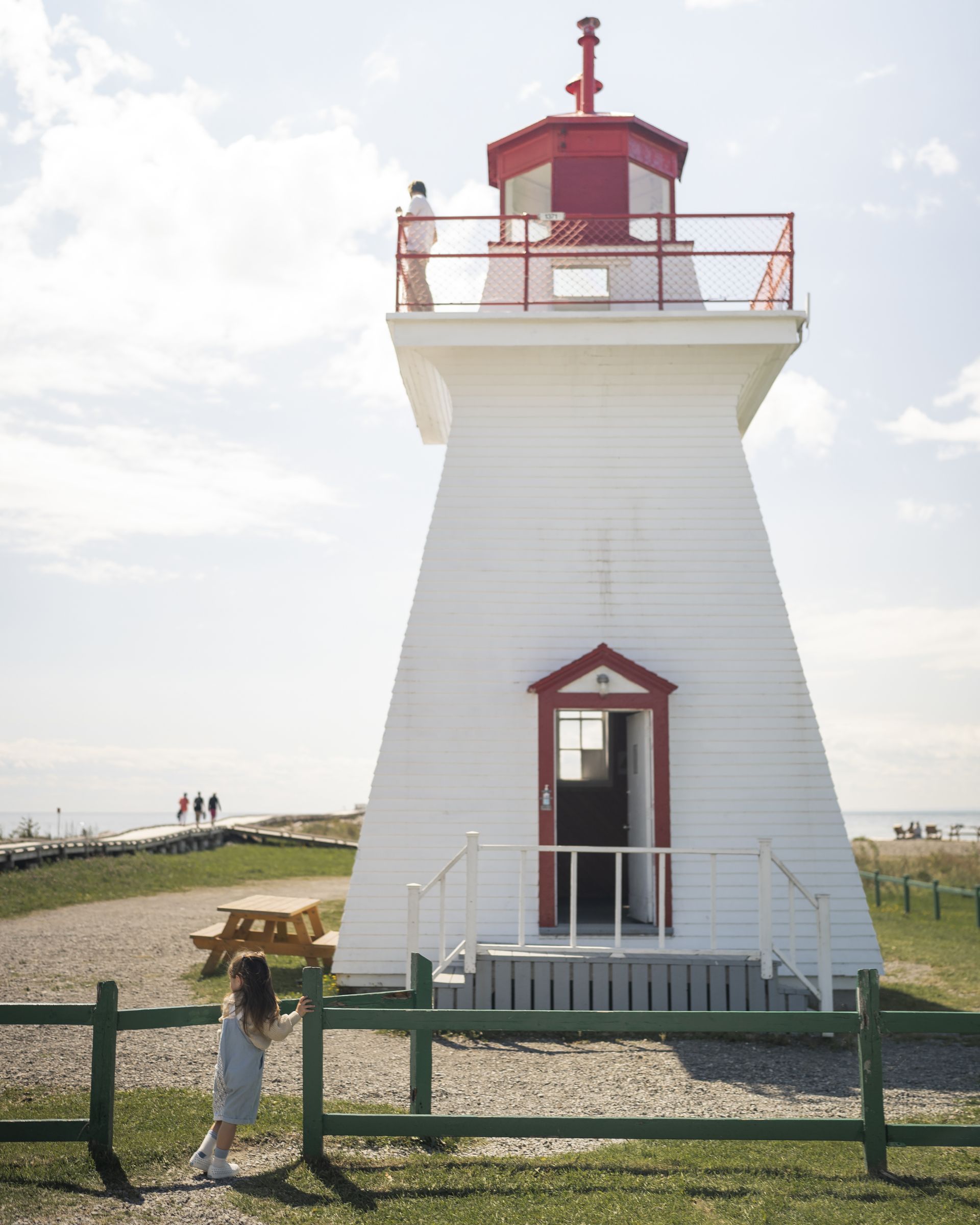 Witte vuurtoren in Canada met rode top en balkon op een grasrijke kust, met een persoon die langs het hek loopt.