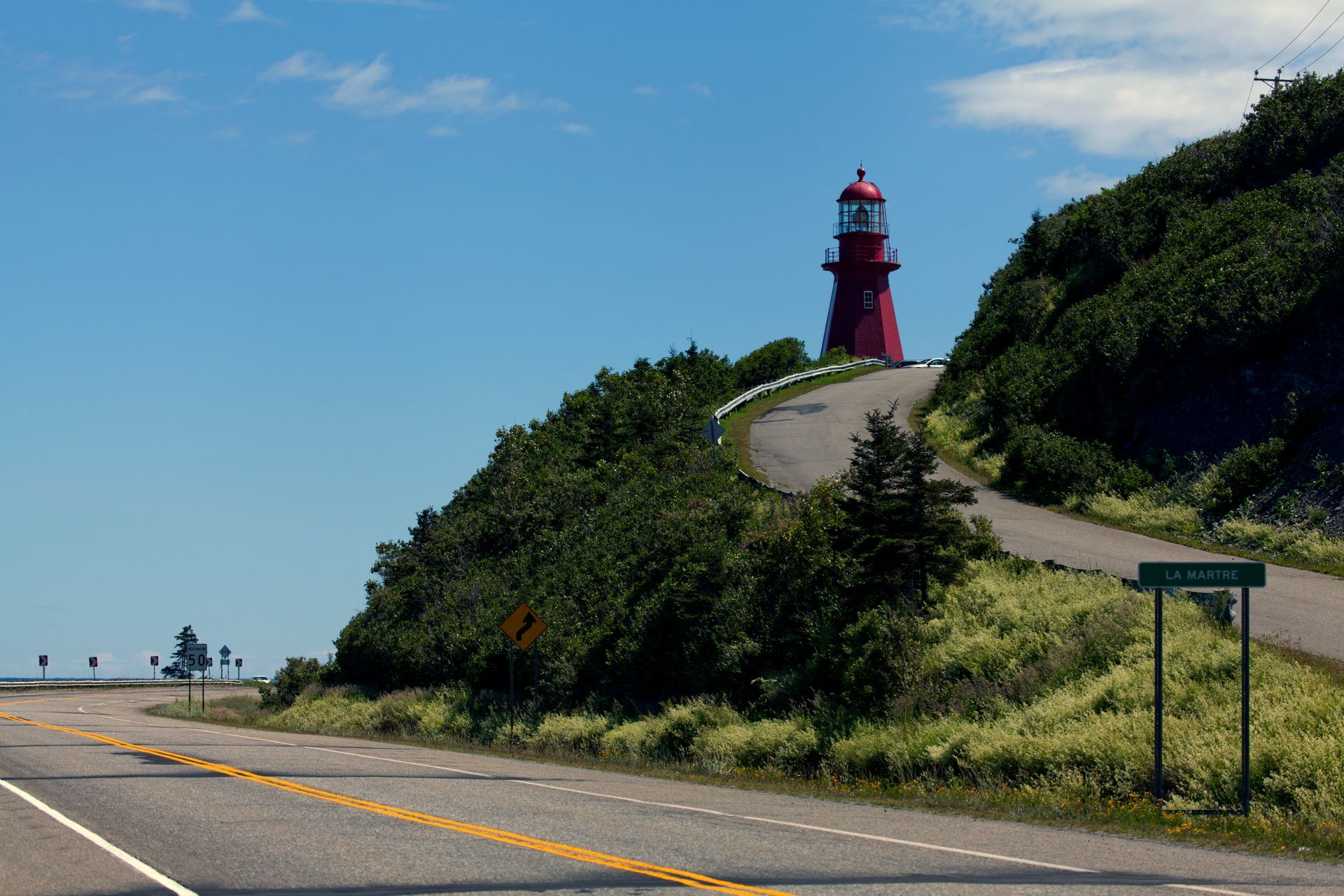 De rode vuurtoren van Le Martre in Quebec, Canada op een grasheuvel langs een kronkelende kustweg onder een blauwe hemel.