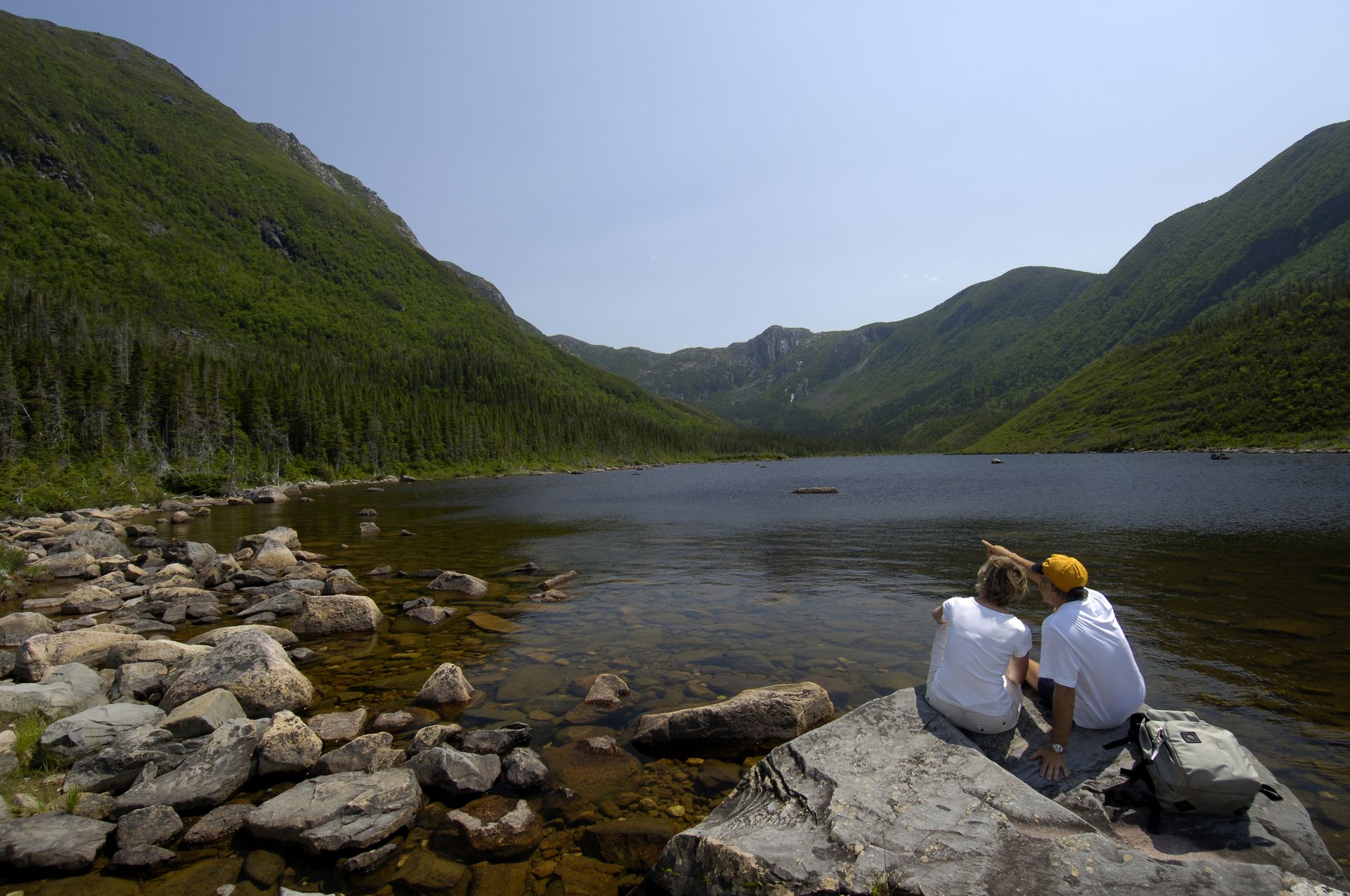 Twee mensen zitten op rotsen aan de rand van het bergmeer Lac aux Américains in het Parc national de la Gaspési in Quebec, Canada, omringd door groene heuvels en kalm water.