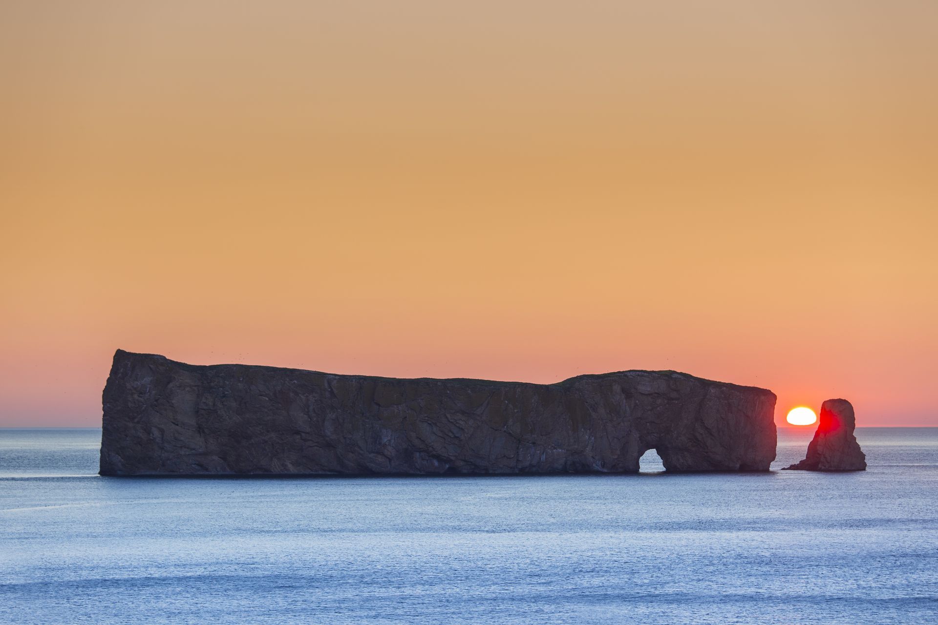 Percé Rock in Quebec, Canada, bij zonsopkomst boven een kalme zee, met de zon die door een gat in de klif schijnt.