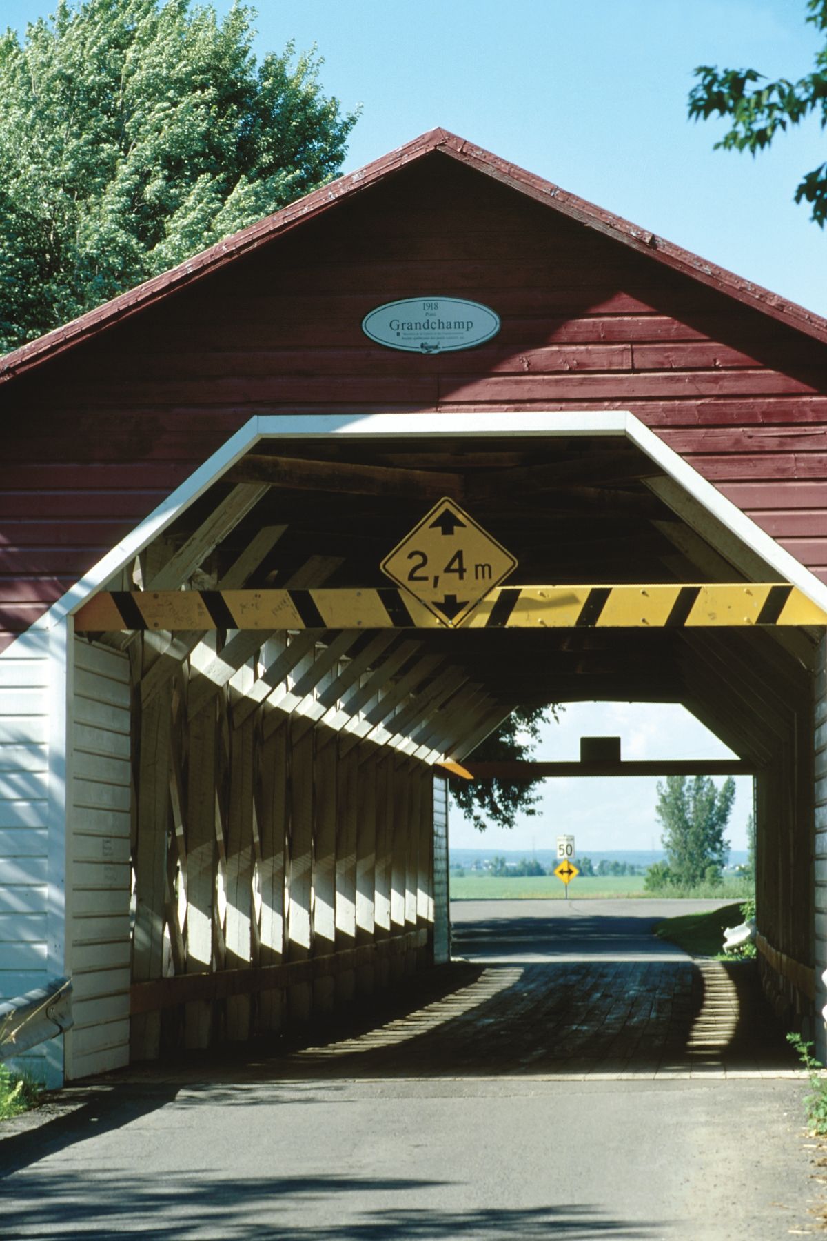 Grandchamp covered bridge in Sainte-Geneviève-de-Berthier, Canada, met een doorrijhoogte van 2,4 meter, aangegeven met een bord, over een verharde weg nabij water.