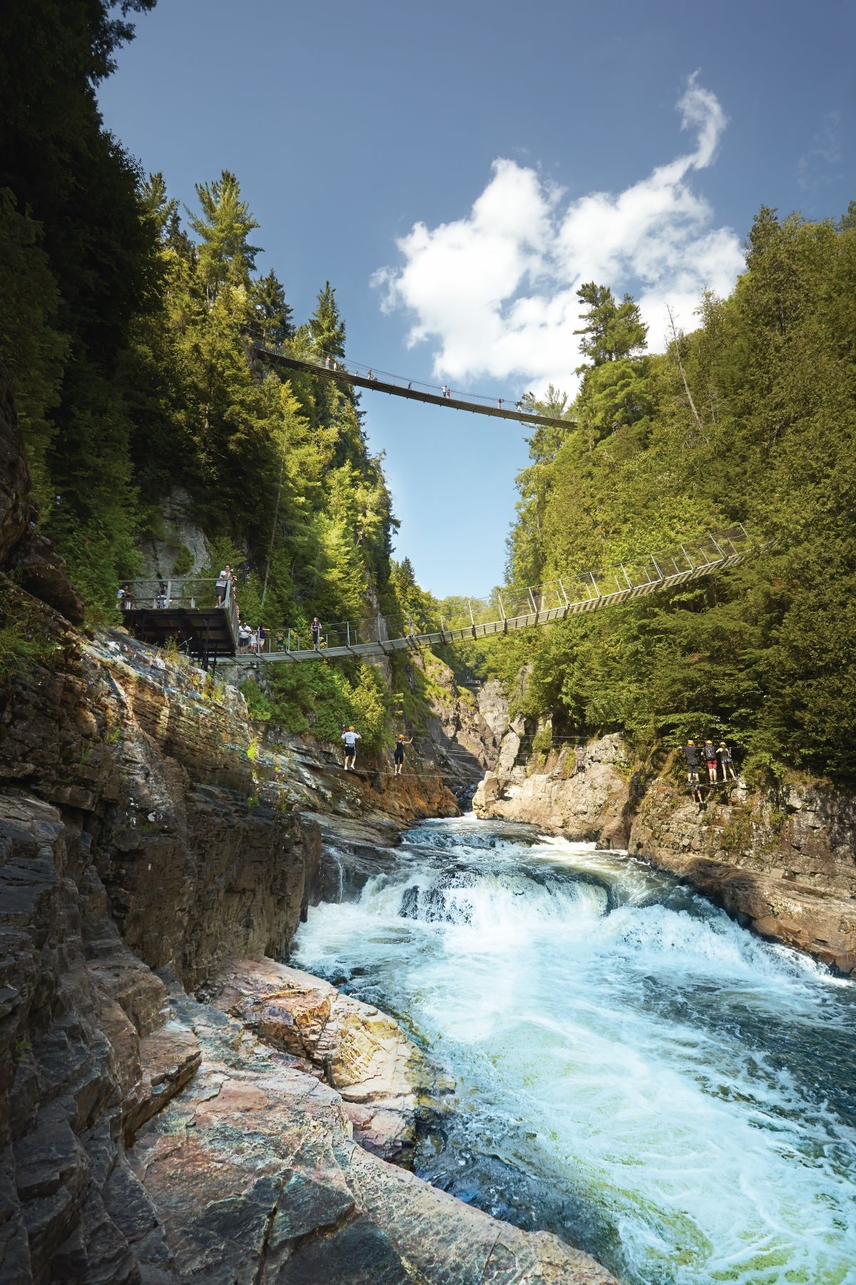 Stroomversnellingen in een bergrivier door de rotsachtige kloof Canyon Sainte-Anne in Canada met weelderige groene bomen en een hangbrug erboven.