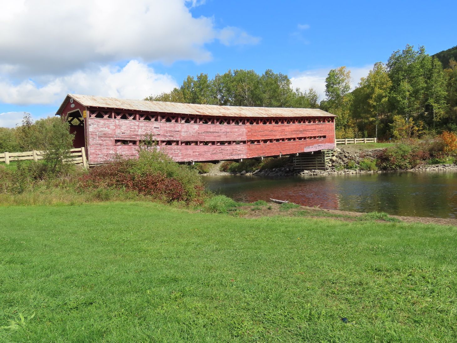 De rode, met hout beklede brug Heppel Covered Bridge in Quebec, Canada, over een rivier naast een grasveld en herfstbomen.