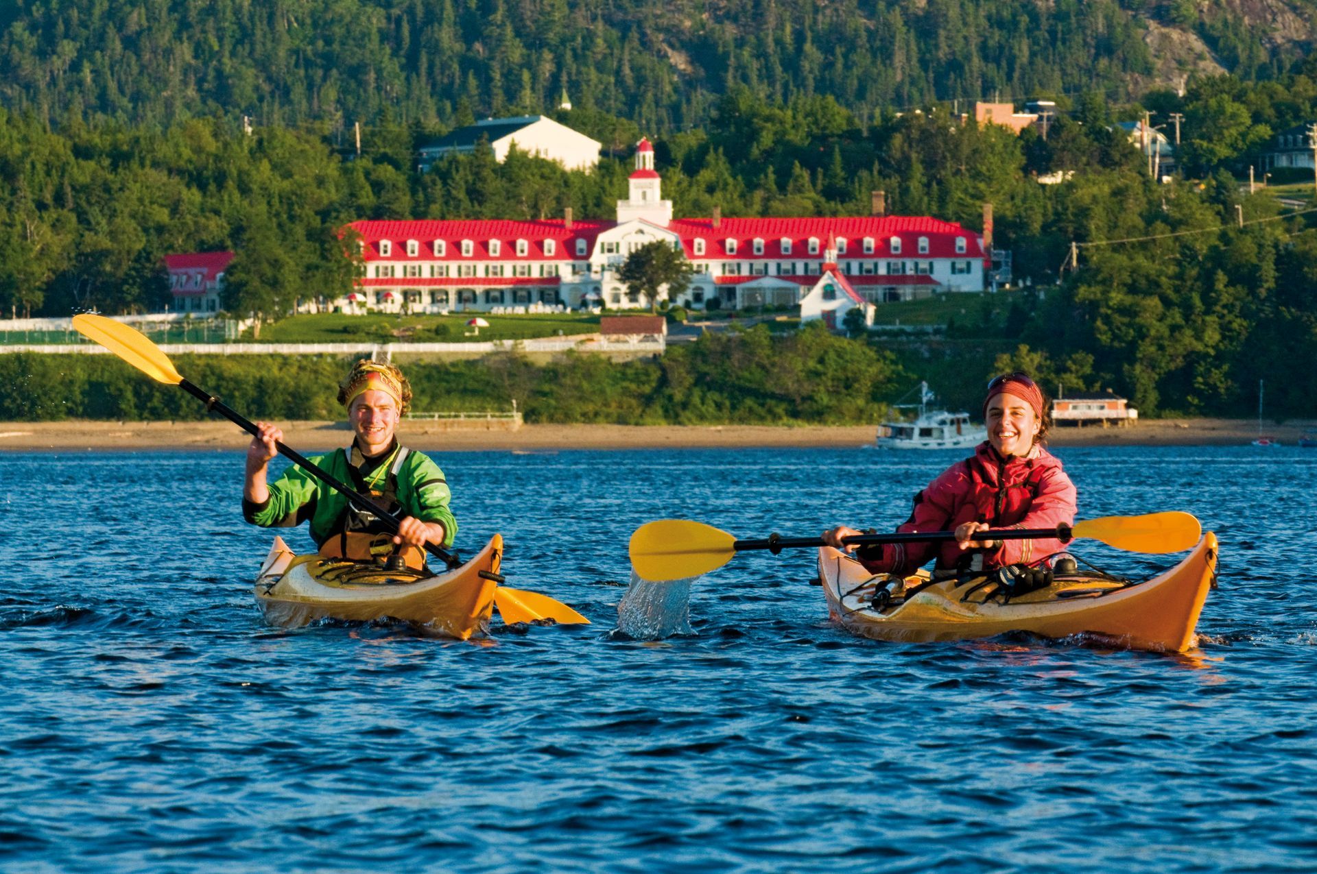 Twee kajakkers peddelen op de rivier met Hotel Tadoussac met een rood dak en beboste heuvels op de achtergrond.