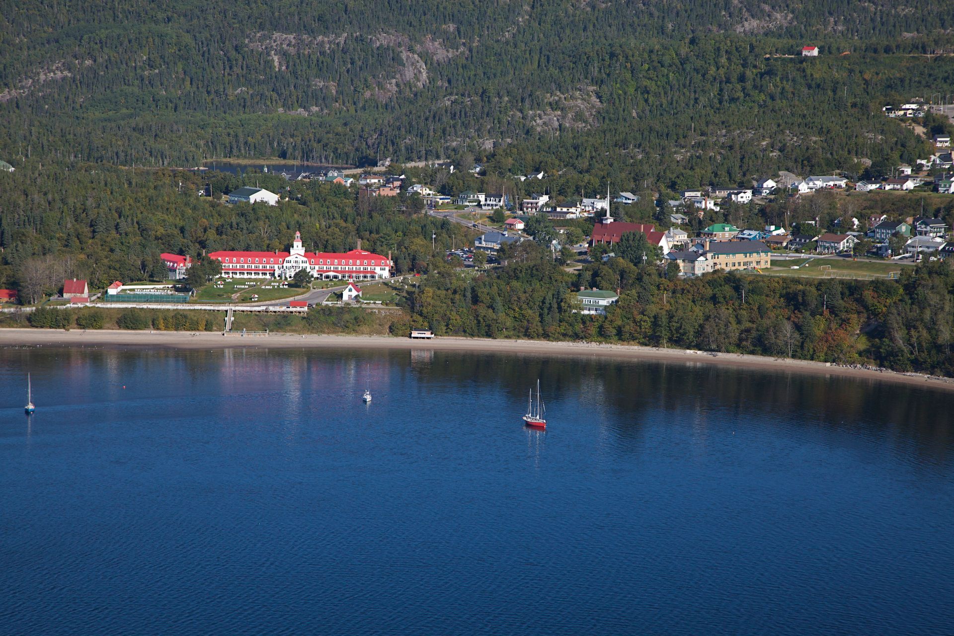 Luchtfoto van Hotel Tadoussac met blauwe baai, kleine bootjes en beboste heuvels op de achtergrond.