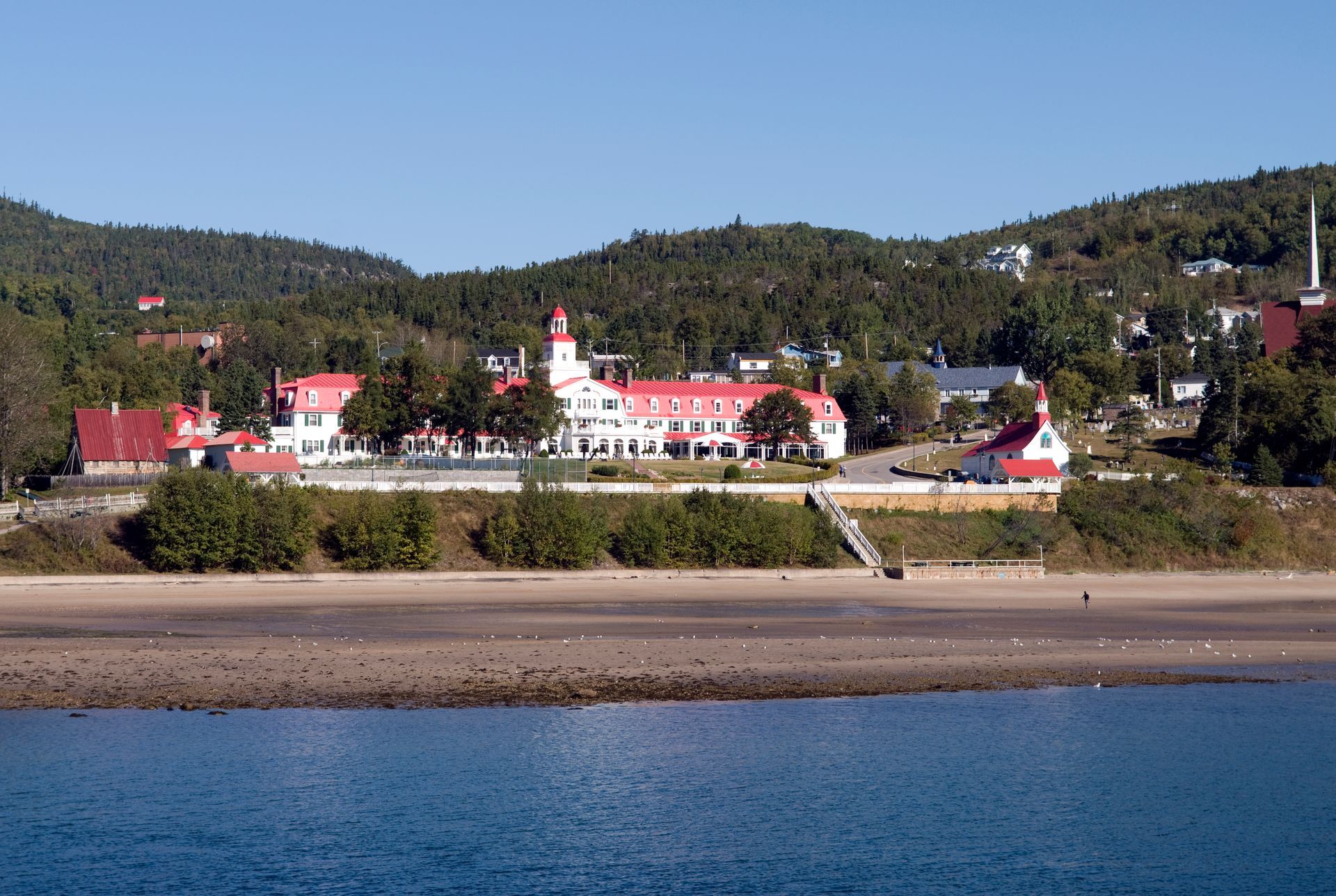 Hotel Tadoussac in Canada met rode daken, witte gebouwen en een kerk, gelegen achter een strand en kalm blauw water onder een heldere hemel.