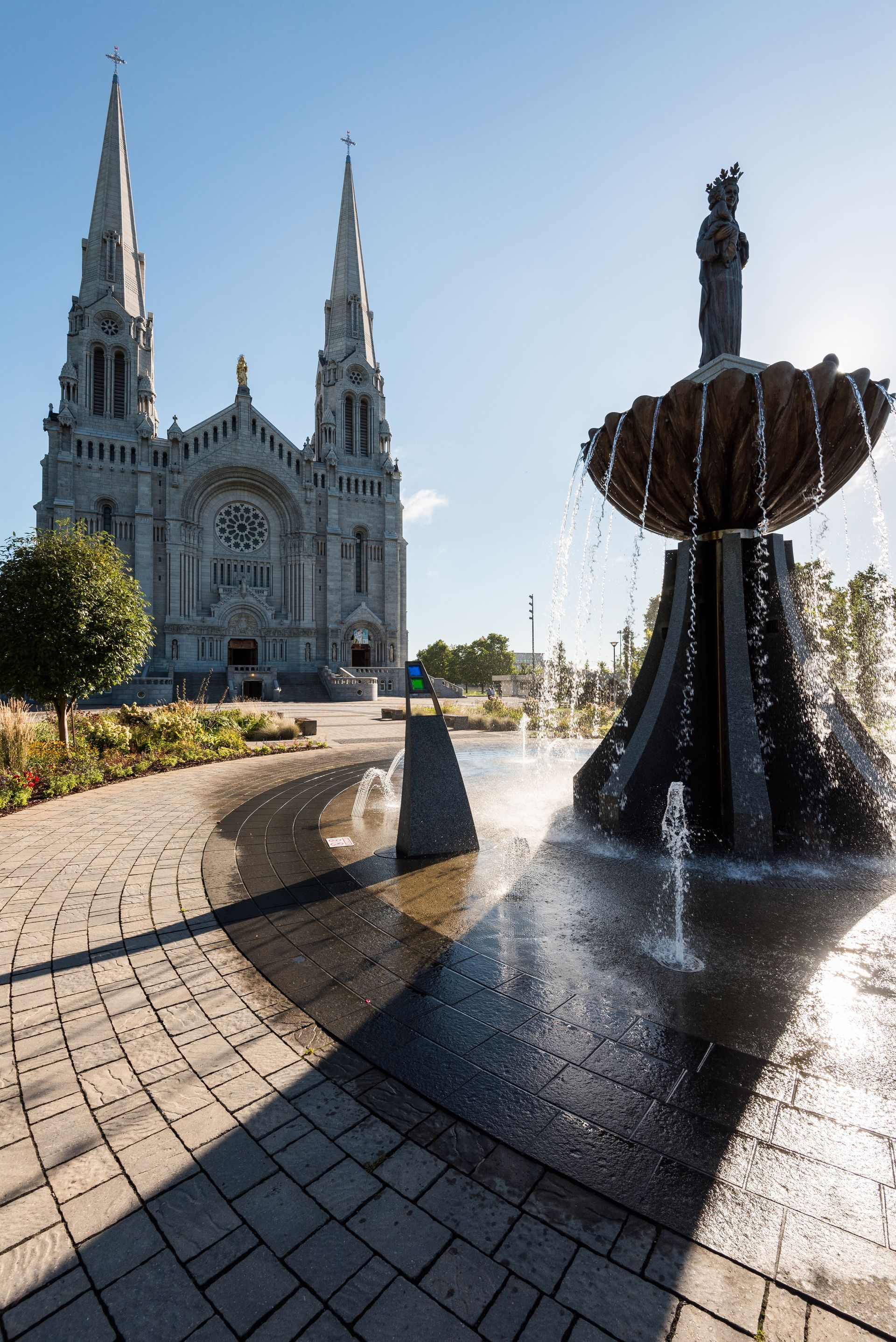 Kathedraalplein rond Sanctuaire Notre-Dame de Sainte-Anne-de-Beaupré in Canada met een sierfontein en een blauwe hemel, met de zon op de voorgrond.