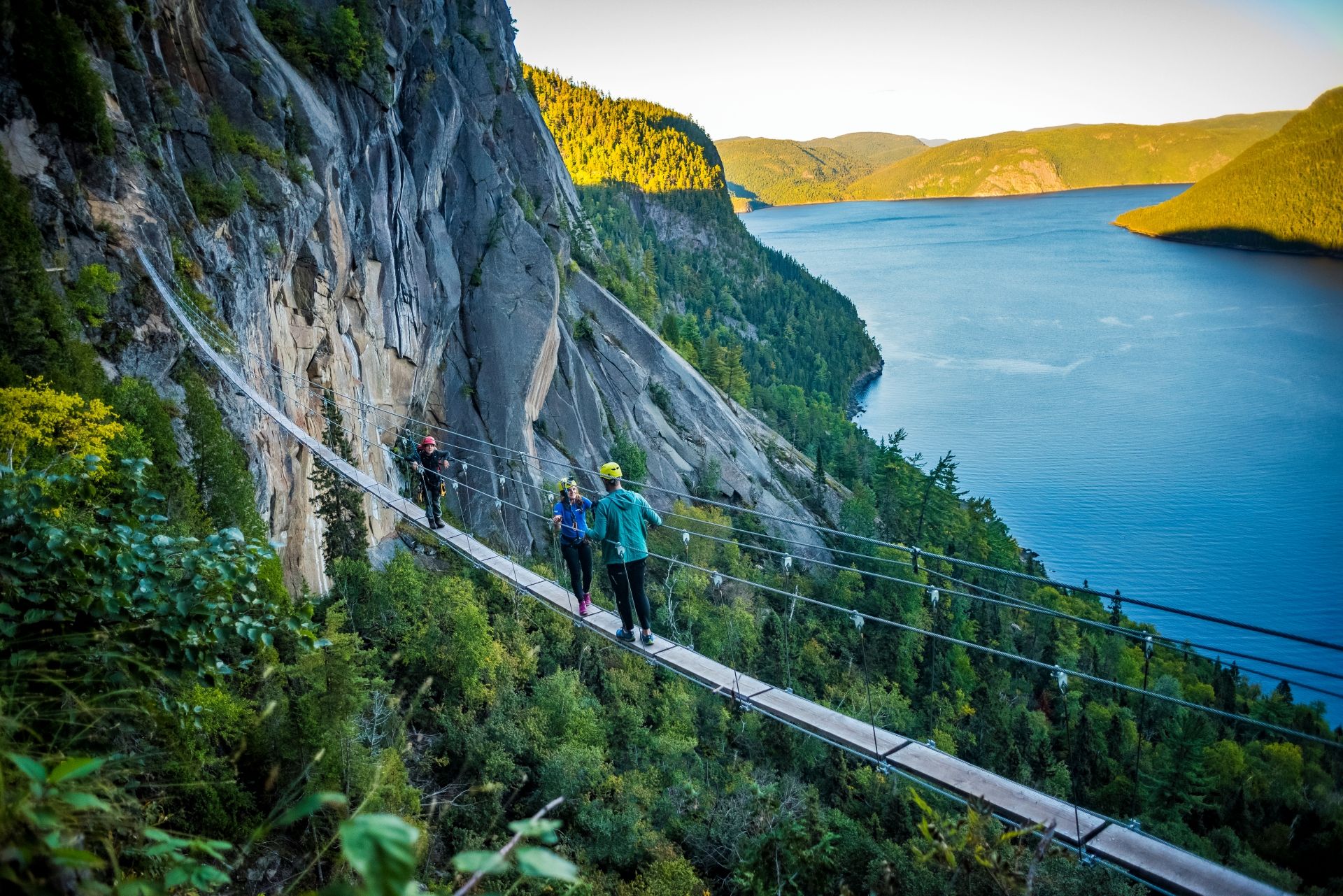 Twee wandelaars op hangbrug langs de klif boven de blauwe Saguenay Fjord in Canada, met steile rotsen en groene heuvels.