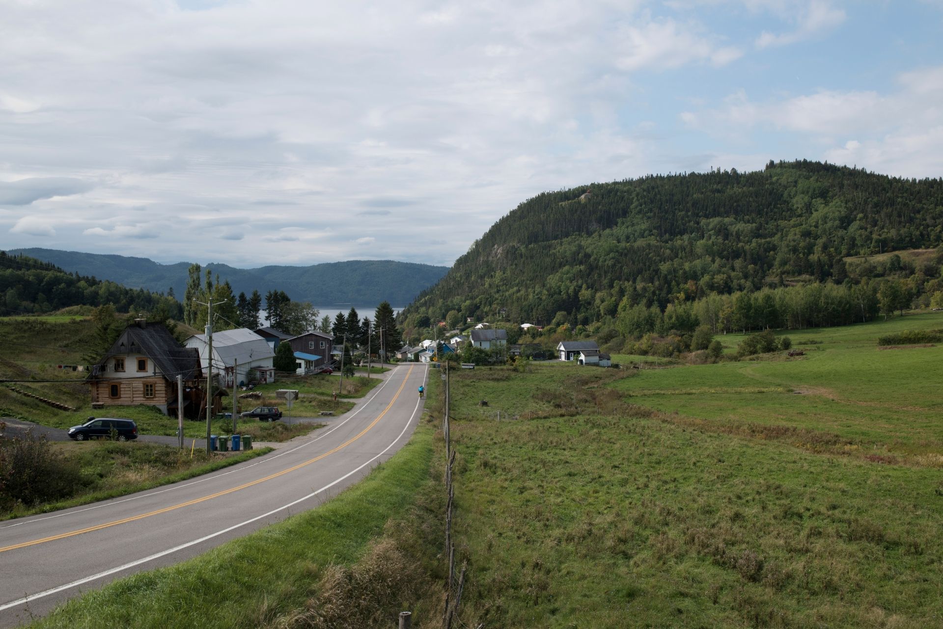 Een landweg slingert door groene velden richting huizen van Sainte-Rose-du-Nord in Canada en beboste heuvels onder een bewolkte hemel.