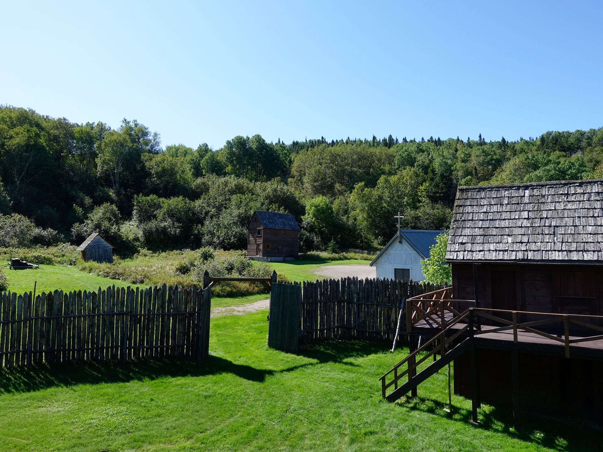 Een zonnige, landelijke tuin van van Val-Jalbert historical Village in Canada met houten, kleine schuurtjes en een grasveld onder een helderblauwe hemel.
