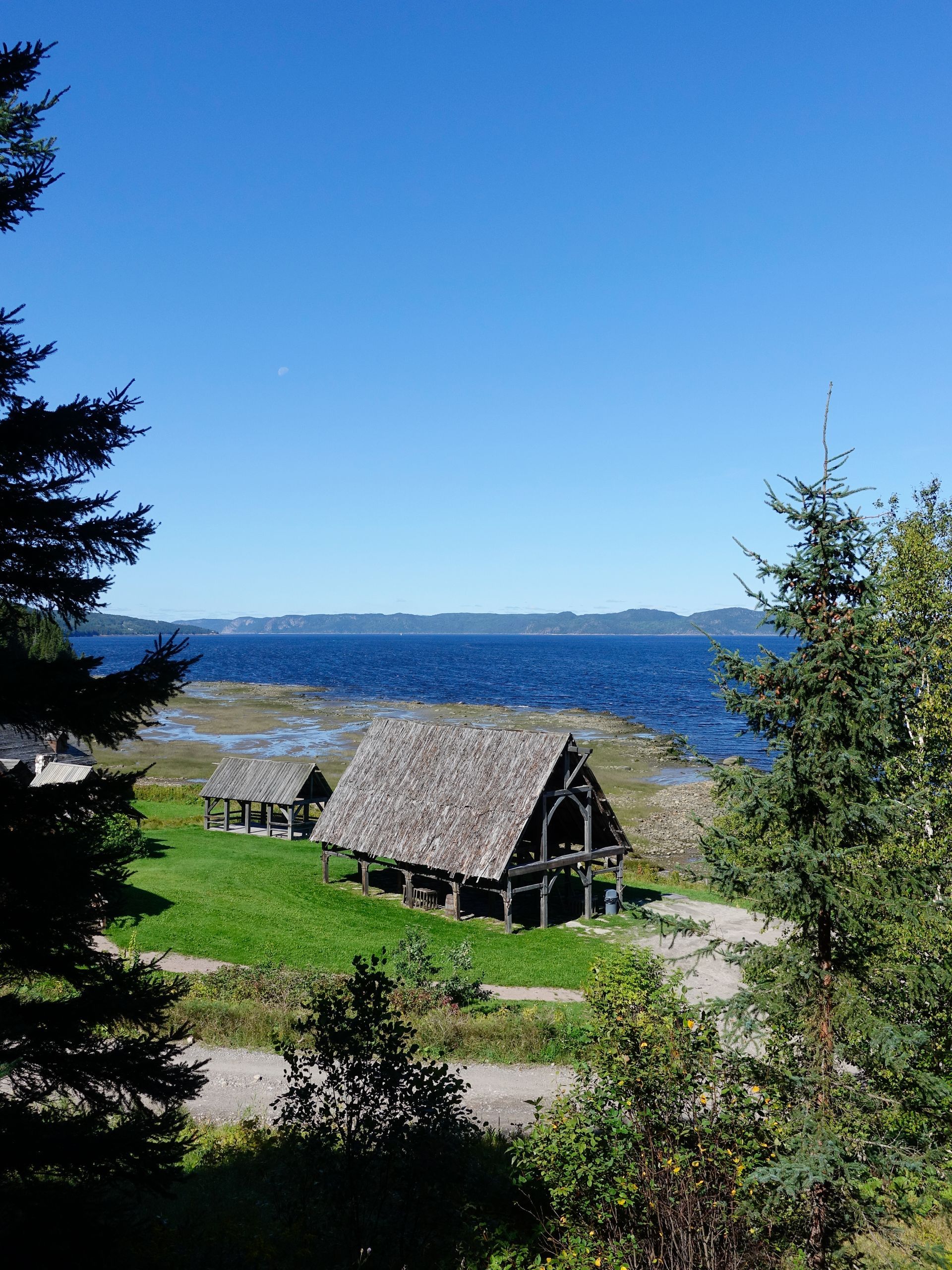 Rustiek huisje van Val-Jalbert historical Village in Canada aan een blauw meer, met groen gras en bomen onder een heldere hemel.