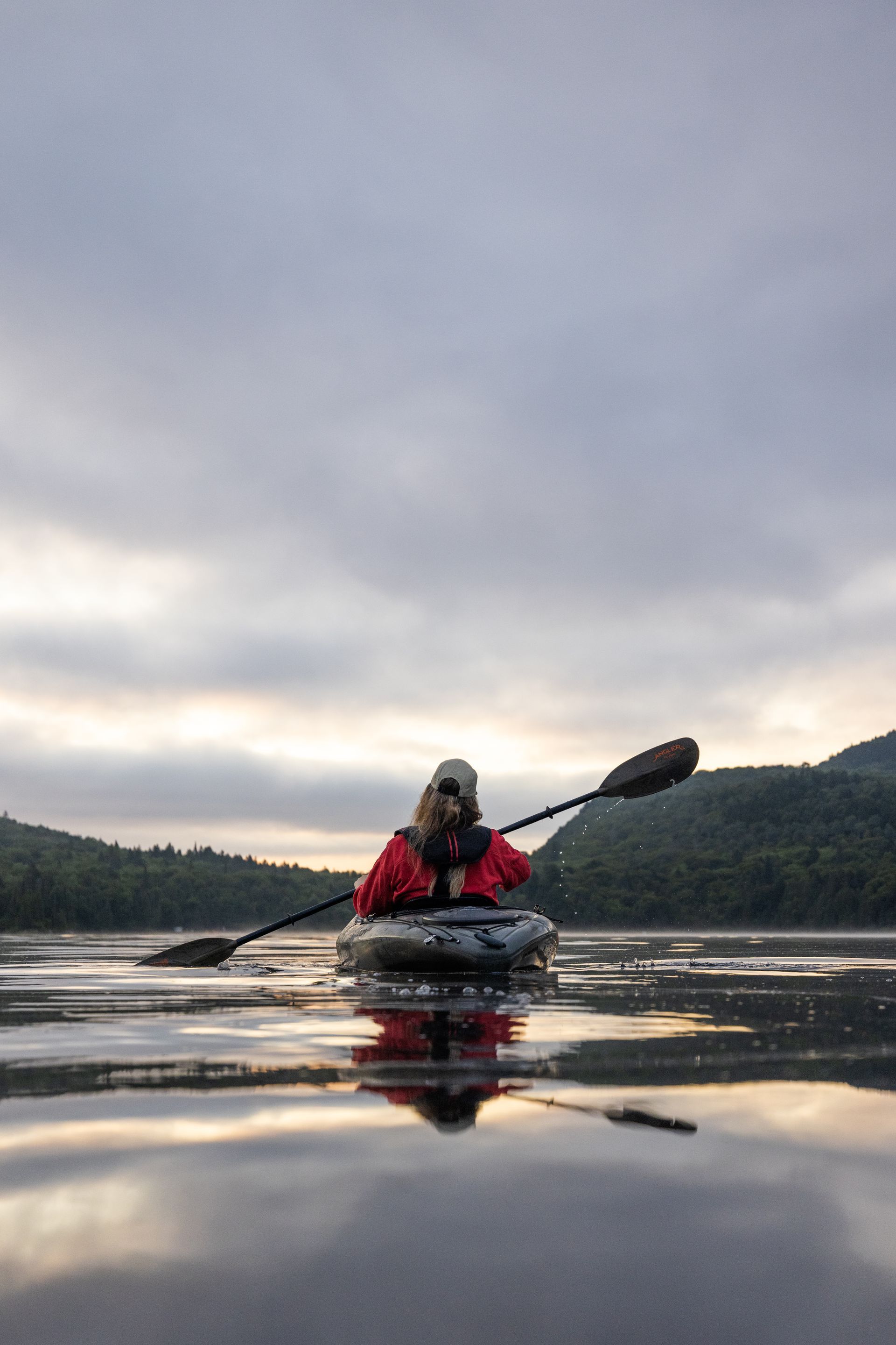 Een kajakker in Mauricie, Canada, in een rode jas peddelt op een kalm meer in de schemering, met bergen en een bewolkte hemel die in het water worden weerspiegeld.