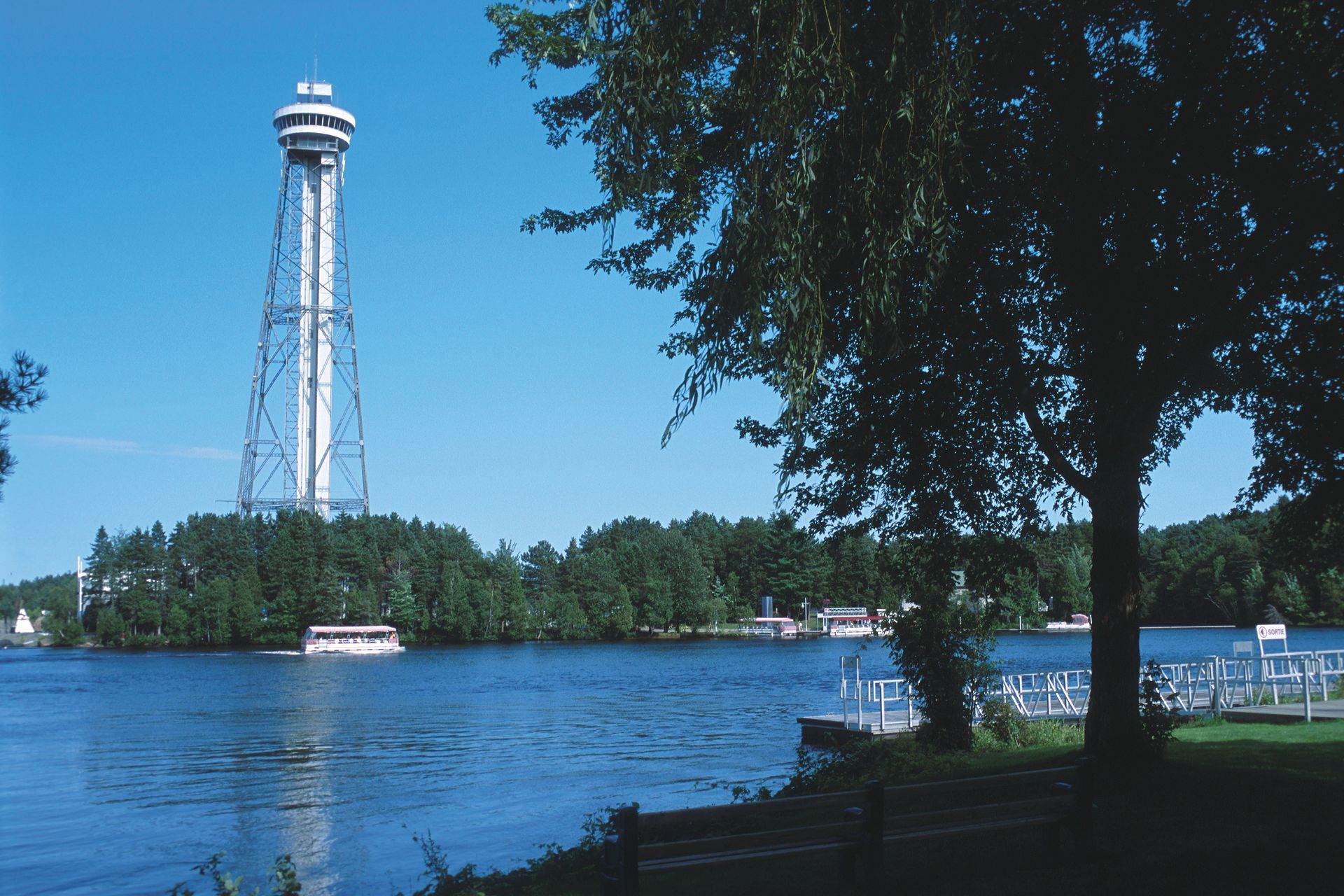 Hoge uitkijktoren in Mauricie, Canada, aan een blauwe rivier met boten en bomen op een zonnige dag.