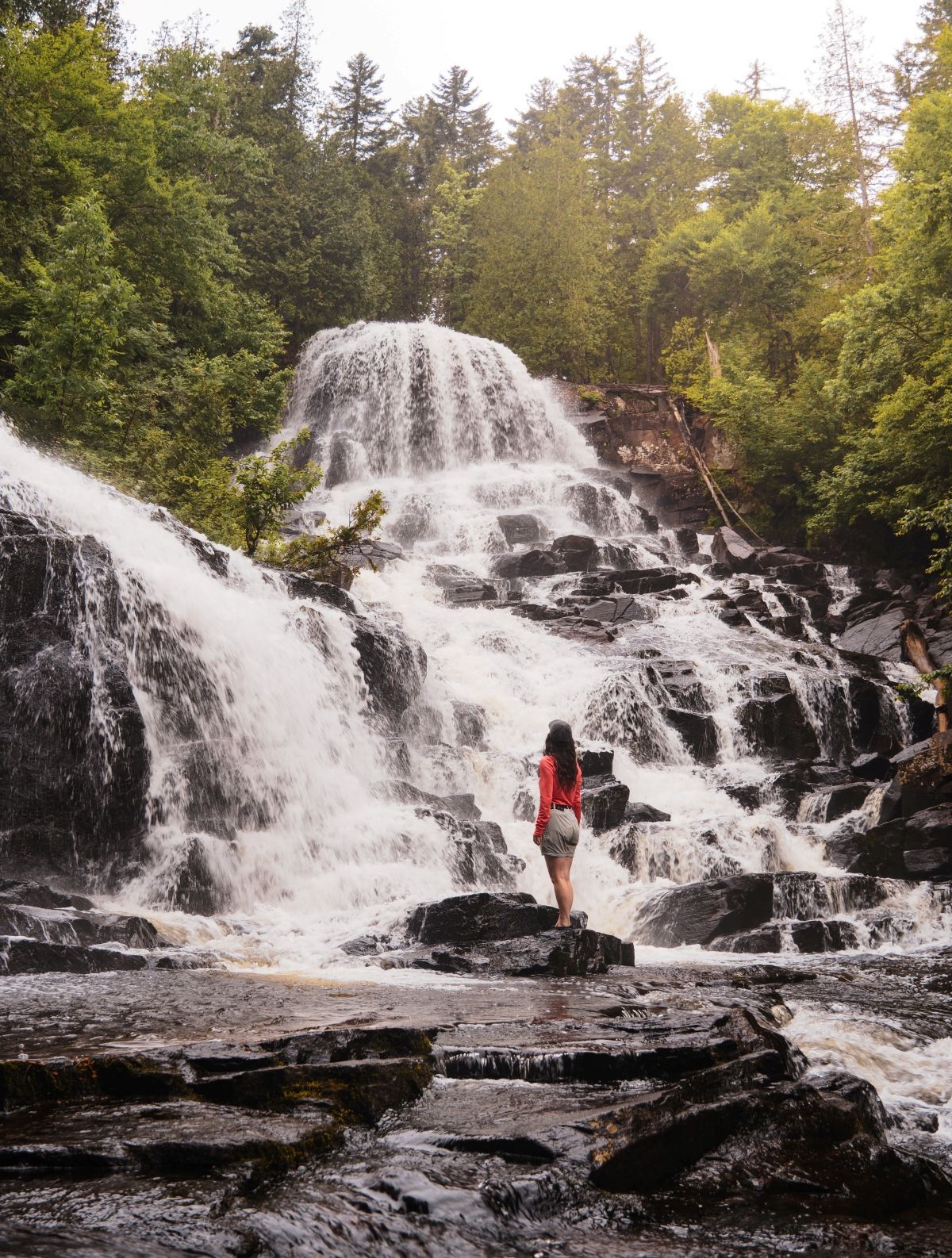 Persoon met rode rugzak staat op rotsen onder de waterval Waber Falls in het La Mauricie National Park in een bos.
