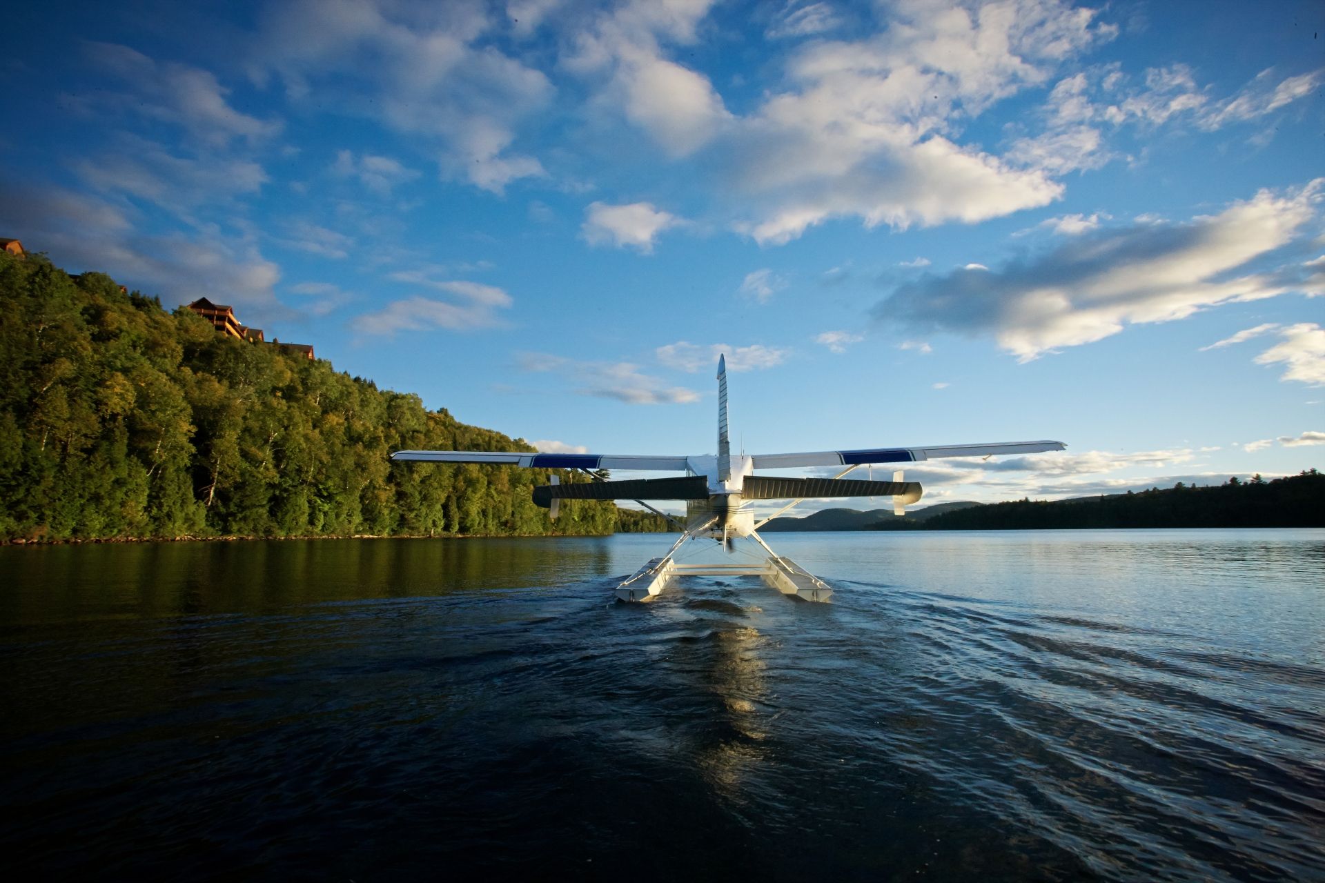 Een watervliegtuig in Mauricie, Canada, scheert over een kalm meer bij zonsondergang, met een beboste oever en een blauwe lucht.