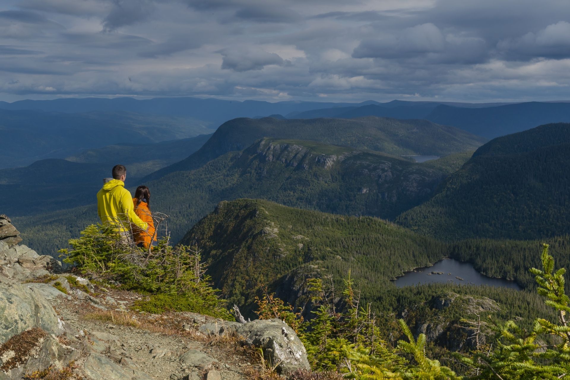 Twee wandelaars in gele en oranje jassen kijken uit op Mont Chic-Chocs in het Parc national de la Gaspésie en een meer onder een bewolkte hemel.