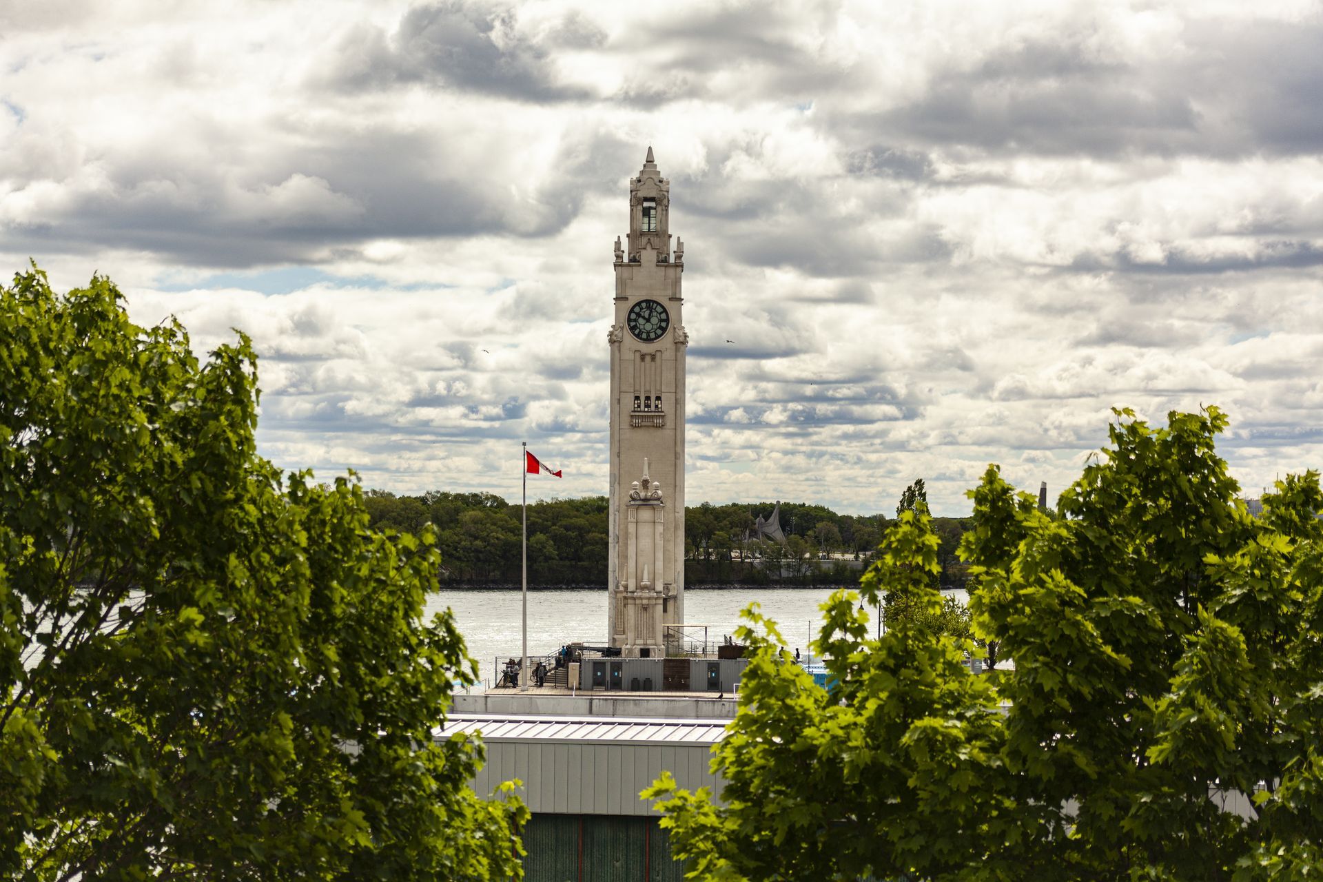 De Clock Tower en de Canadese vlag staan naast een rivier in Montreal, omringd door groene bomen onder een bewolkte hemel.