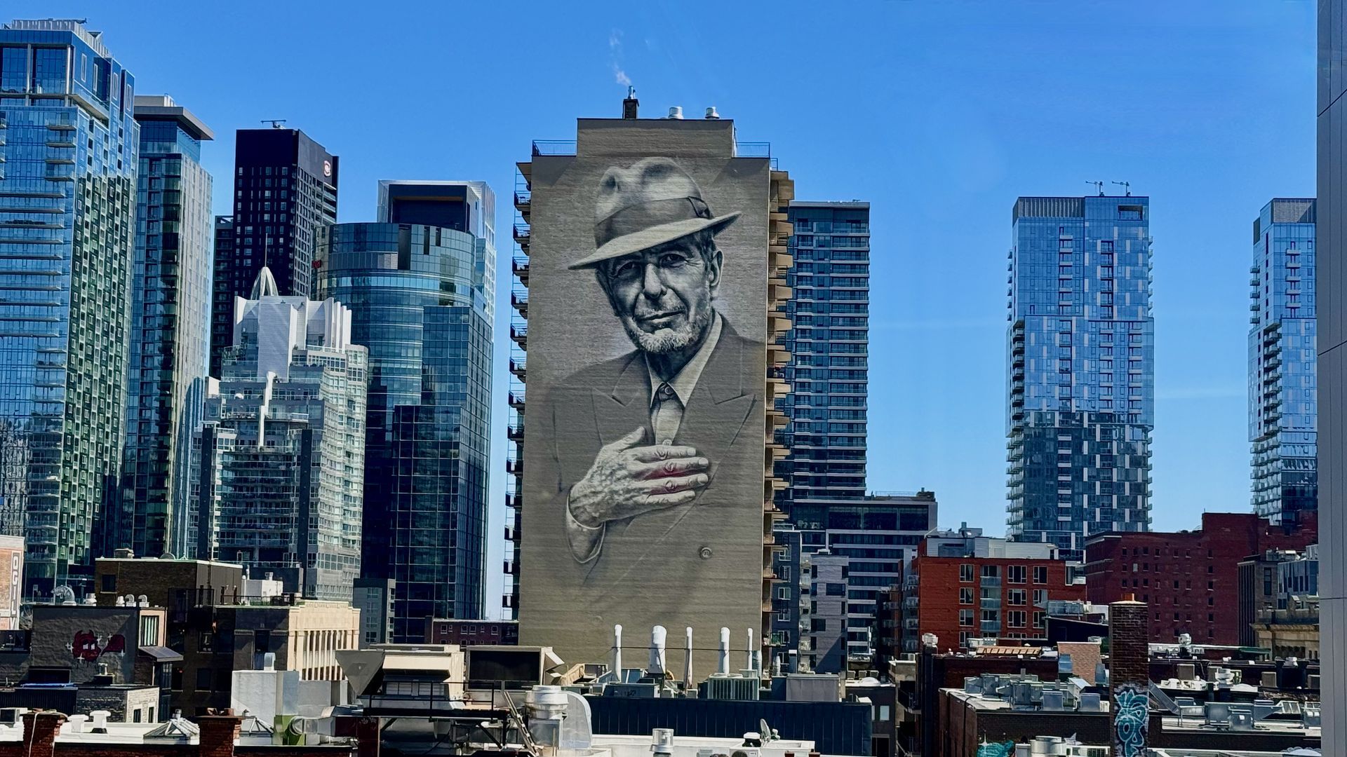 De skyline van Montréal met een grote muurschildering Leonard Cohen met een hoed op een hoog gebouw, onder een helderblauwe hemel.
