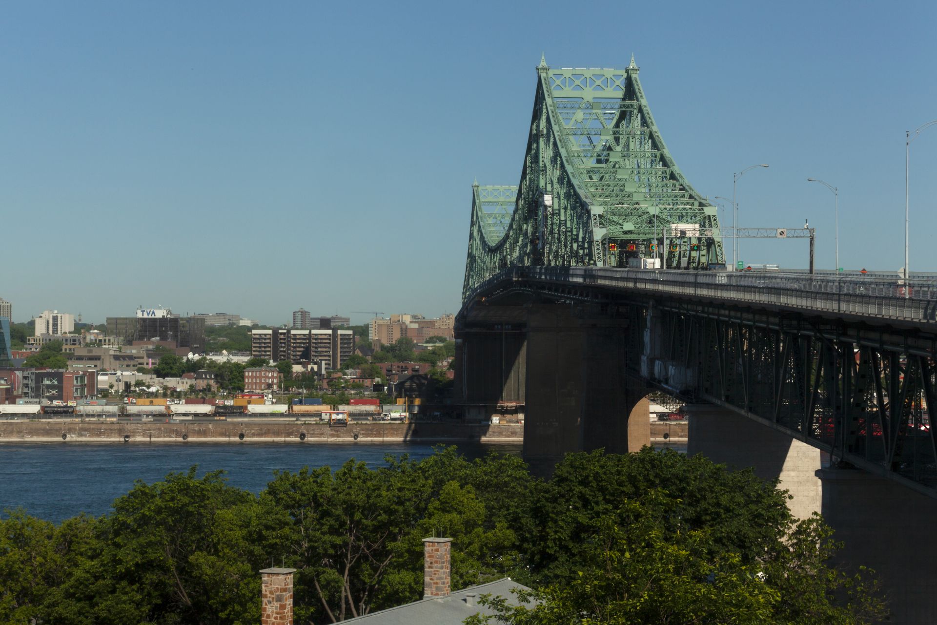 Stalen brug in Montreal over een rivier met een groene vakwerktoren en de skyline van de stad op de achtergrond.