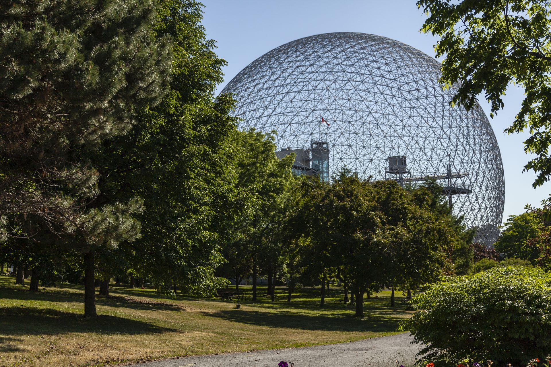 Een geodetische koepel in een park in Montreal, gedeeltelijk verscholen achter bomen, onder een helderblauwe hemel.