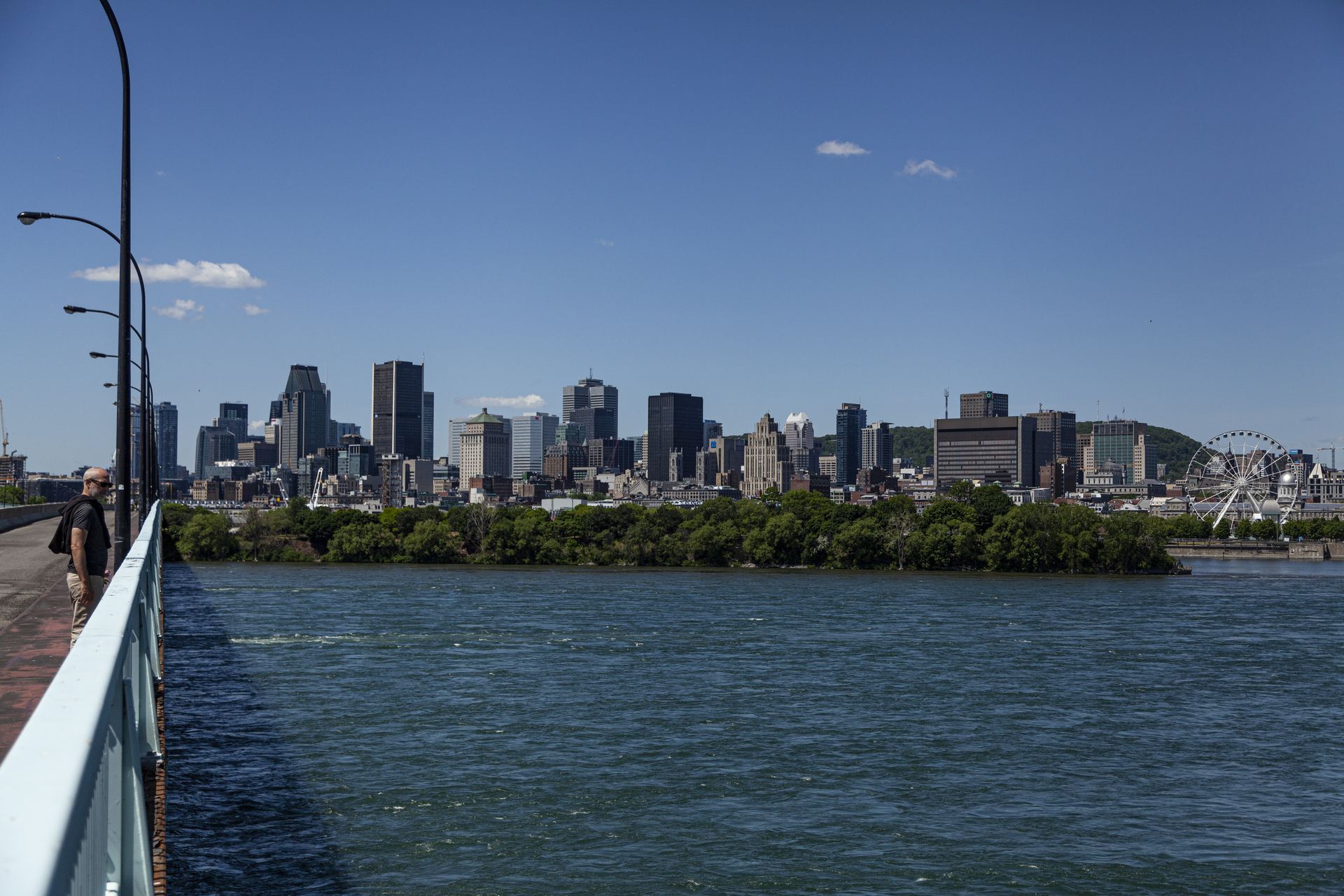 De skyline van Montreal aan de overkant van het water onder een blauwe hemel, gezien vanaf een loopbrug.