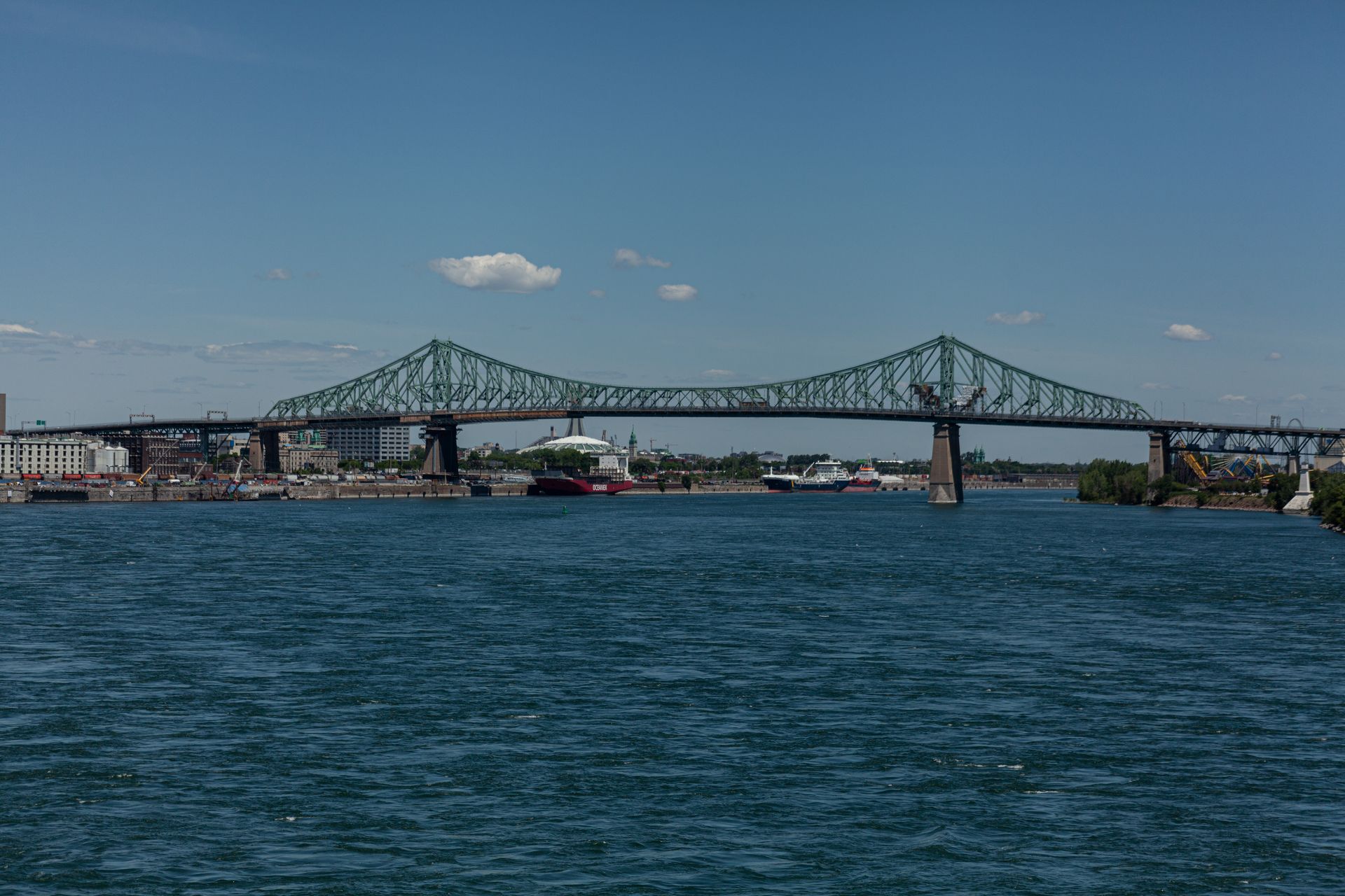 De Jacques-Cartier brug in Montreal overspant een brede rivier onder een helderblauwe hemel, met kabbelend water op de voorgrond.