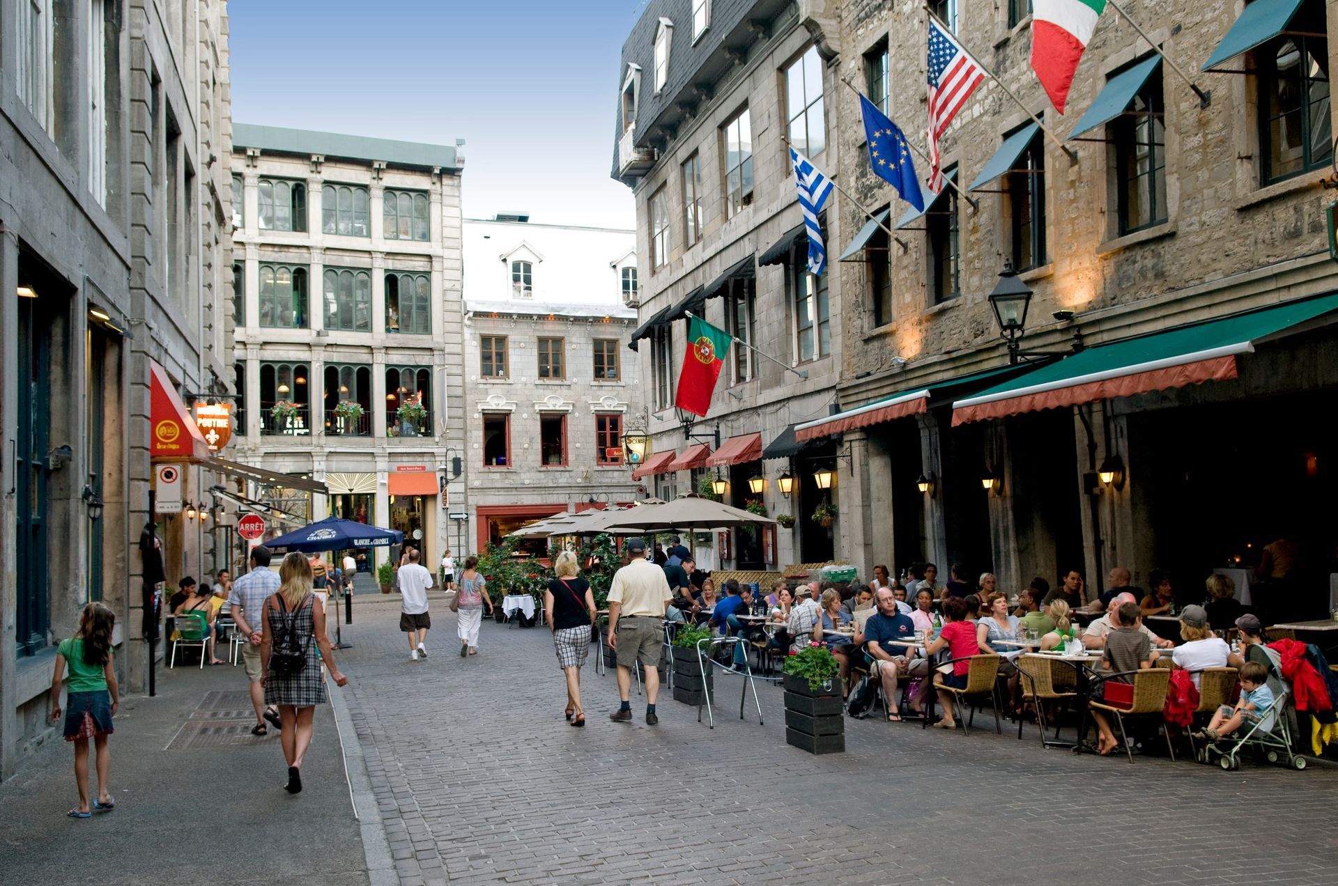 Een drukke straat in Montreal met kinderkopjes, waar mensen wandelen en terrasjes te vinden zijn tussen hoge gebouwen en vlaggen.
